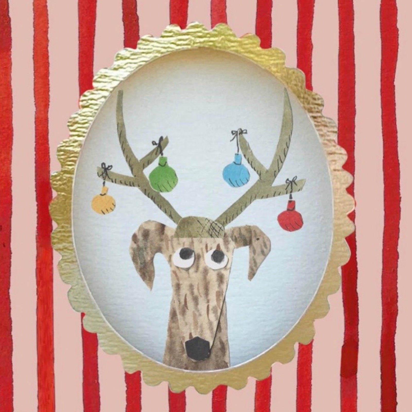 A close up of a card featuring a dog wearing a deer hat with antlers decorated with colourful hanging ornaments, set against a red and pink vertically striped background with a gold scalloped border.