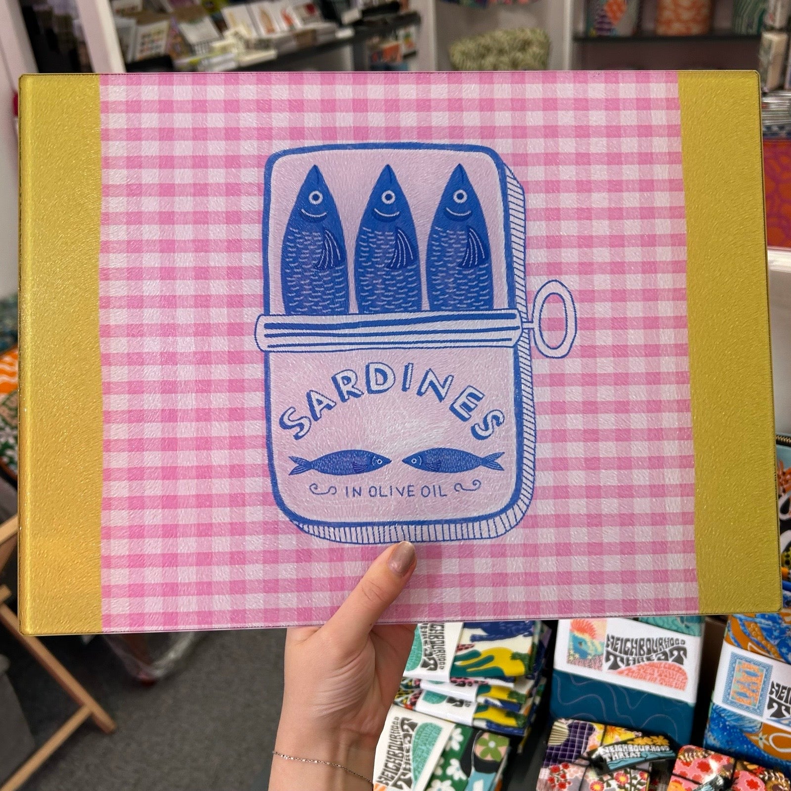 A hand holds a pink and white gingham-patterned chopping board with yellow side panels. The front features a blue illustration of three sardines in a tin labeled 'SARDINES IN OLIVE OIL'.