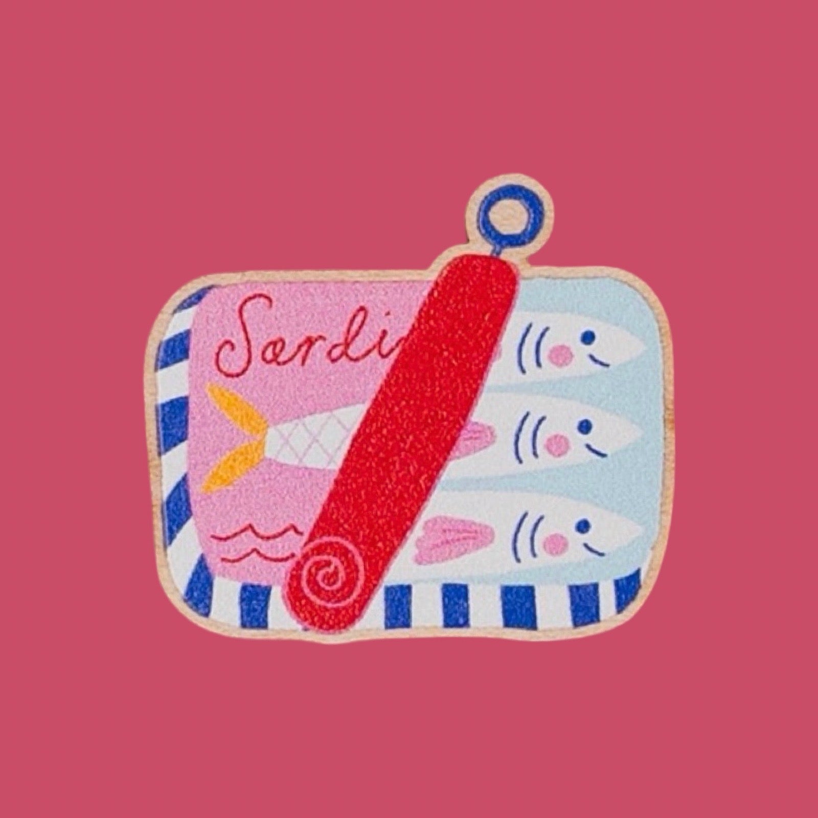 A colourful pin badge shaped like a sardine tin, featuring three cartoon fish on the right, the word 'Sardine' on the left, blue and white stripes around the edge, and a red diagonal band across the centre, shown on a solid pink background.