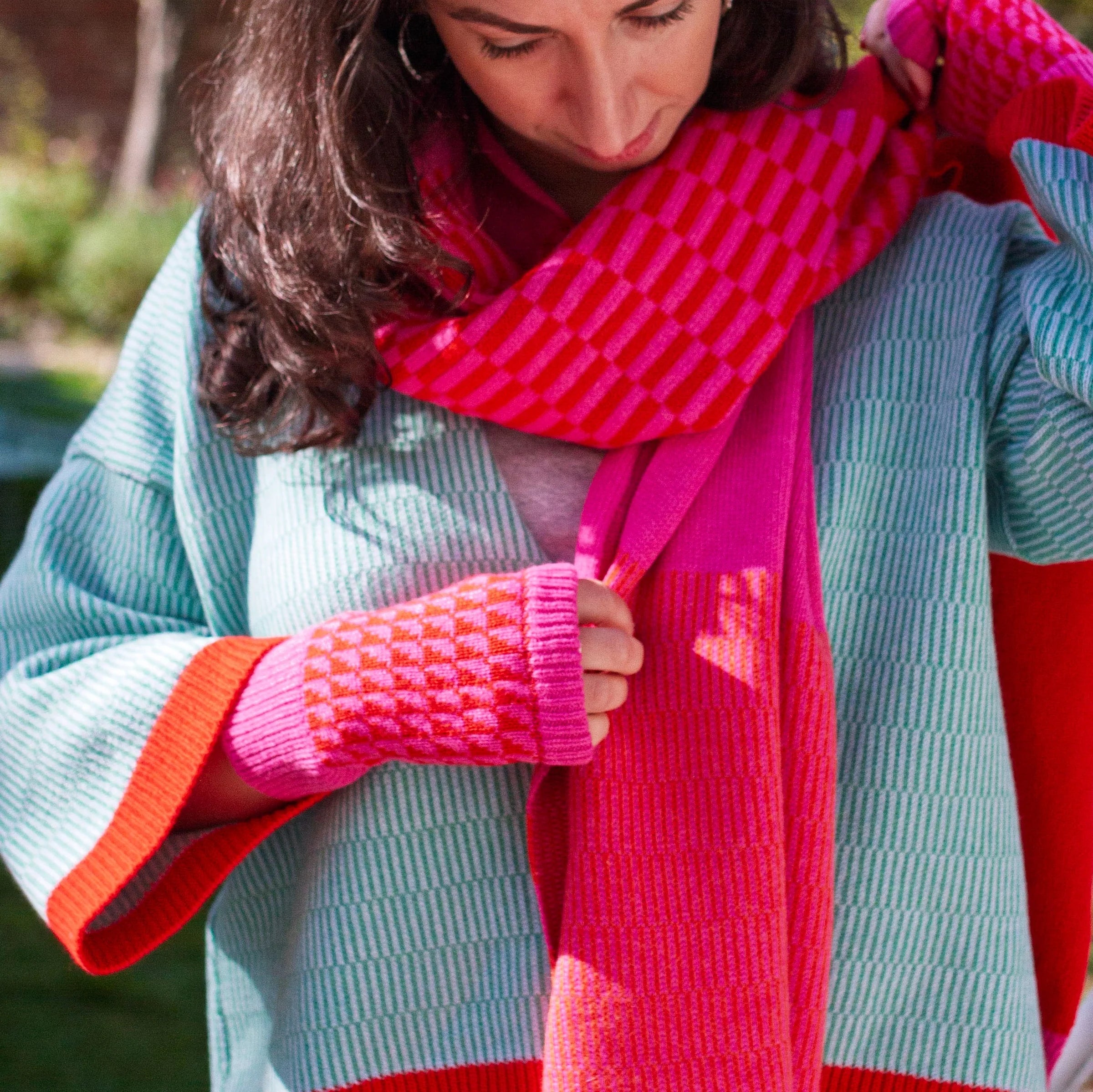A woman outdoors wearing a chunky knit sweater in light blue, paired with a bright pink and red striped scarf and matching pink knit wrist warmers.