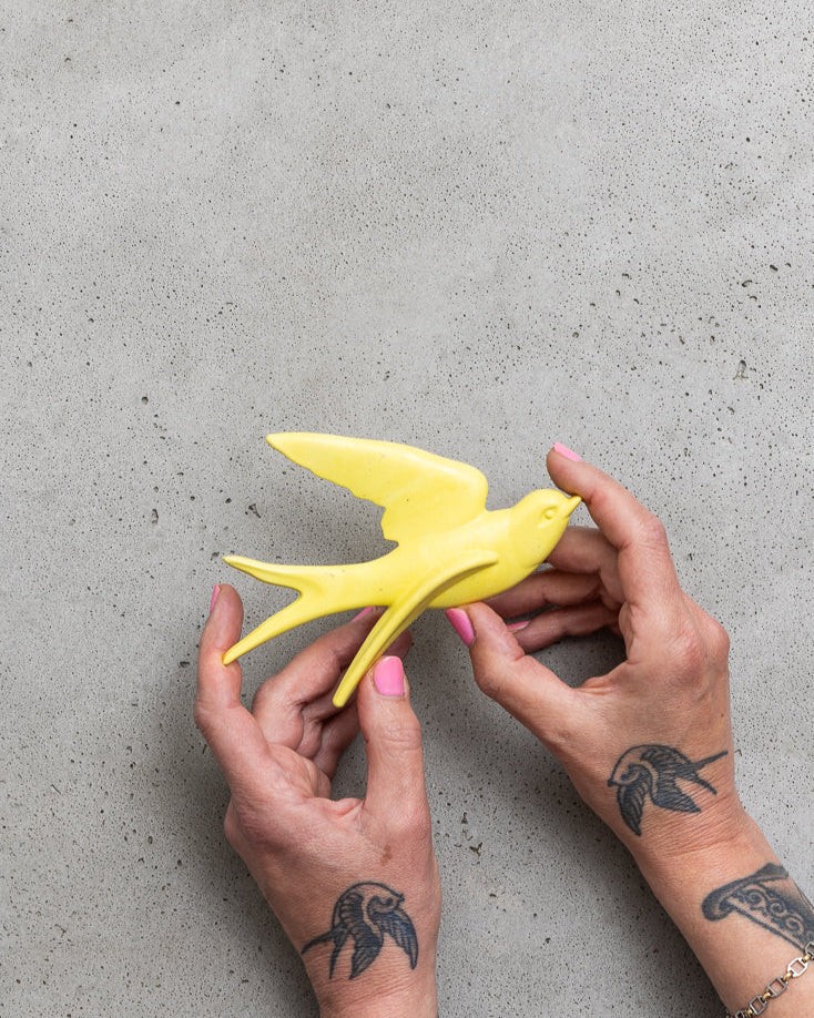 Yellow bird figurine held by hands with tattoos on a grey surface.