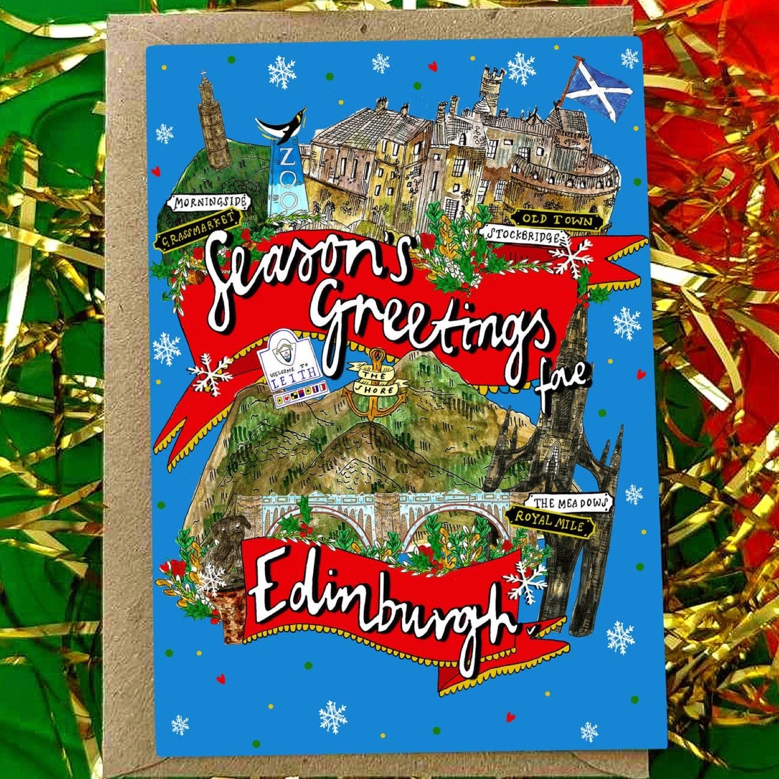 An illustrated greeting card featuring a colourful Edinburgh holiday scene with the text 'Season's Greetings' and 'Edinburgh', placed on a kraft envelope against a festive background of gold and red tinsel.
