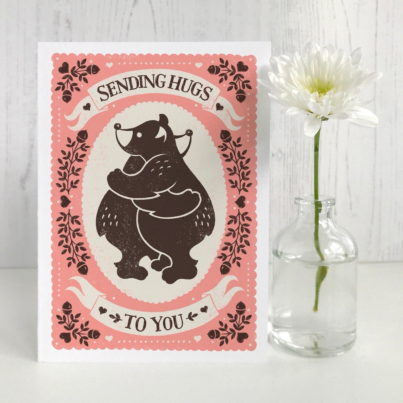 Card with bear illustration and text 'Sending Hugs to You' next to a vase with a flower on a light wooden background.