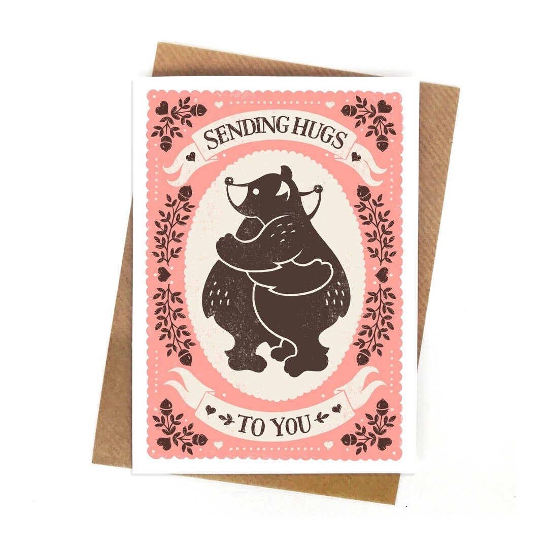 Card featuring two brown bears hugging inside a cream oval in the centre of the card with the text 'sending hugs to you' written in brown text inside white banners above and below the bears, All on a pale pink background with brwon acorns and leaves.