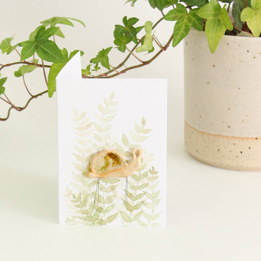 A snail shaped ceramic plant hugger decoration decorated with brown and white recycled glass on a backing card