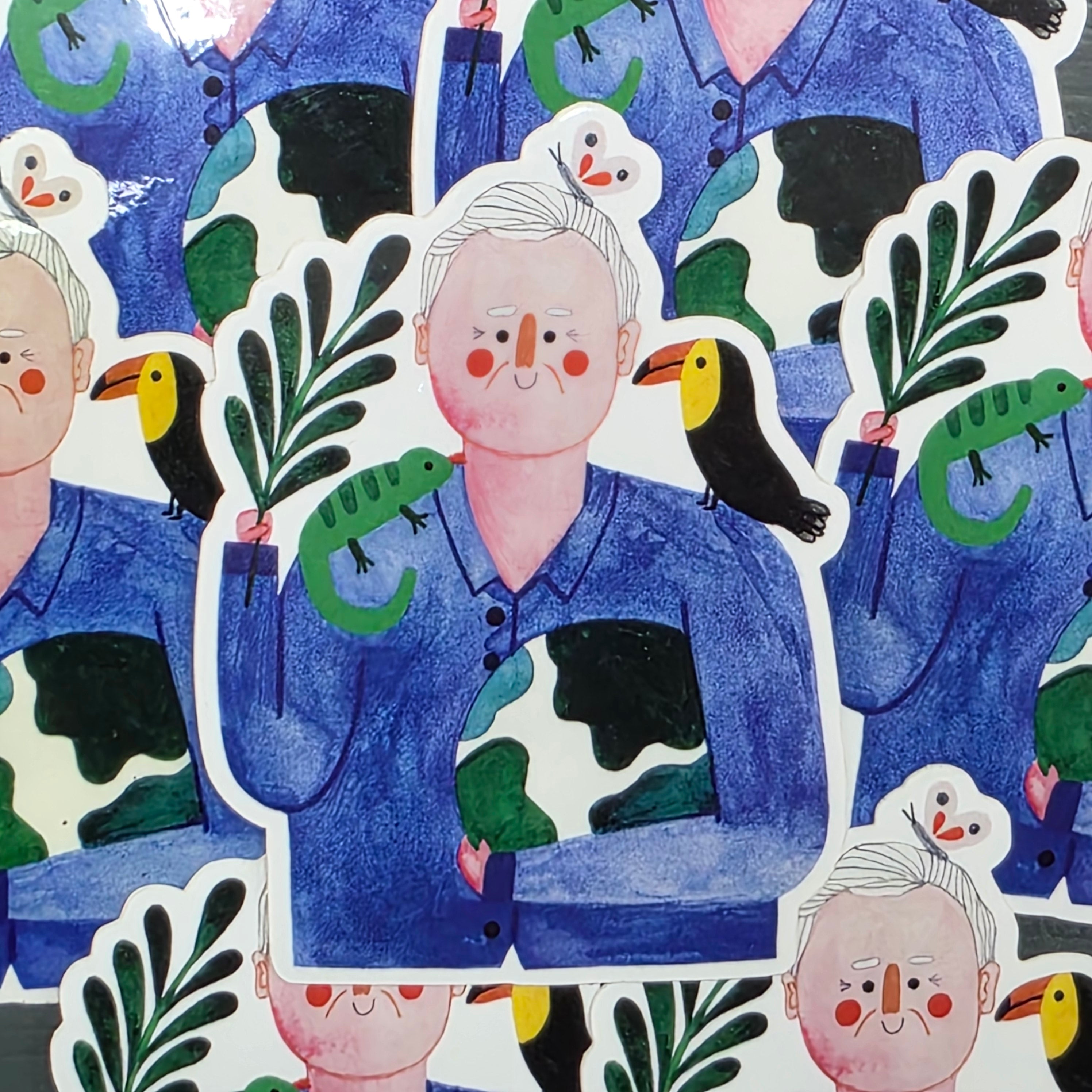 This vinyl sticker features an illustration of  Sir David Attenborough holding a fern in one hand and hugging a globe with the other. He has a toucan on one shoulder and a chameleon on the other.