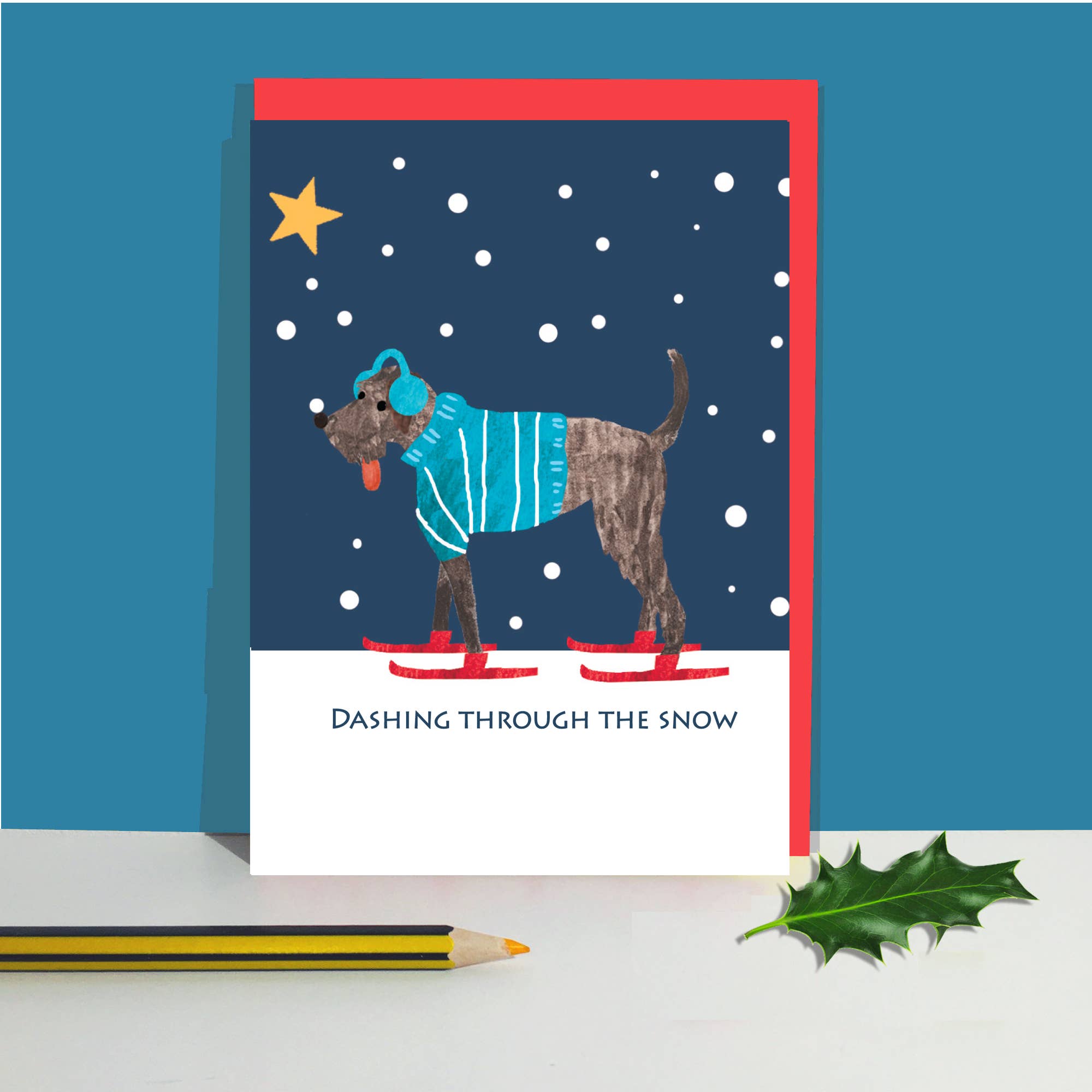 A greeting card featuring an illustration of a dog wearing a blue striped sweater and red skates, with the text 'Dashing Through the Snow' on a white lower section. The card is shown standing upright on a white surface against a blue background, with a pencil and holly leaf.