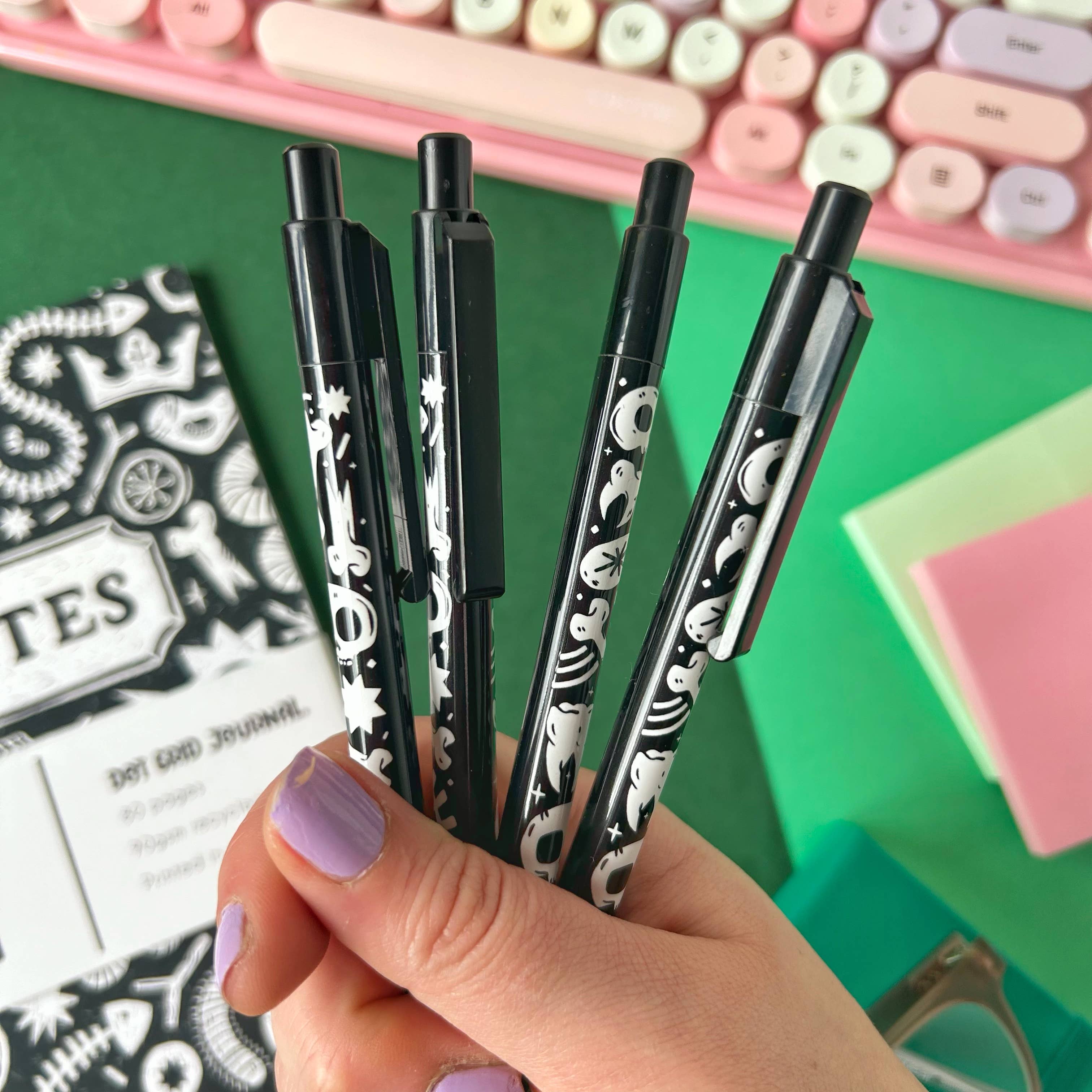 Hand holding black and white skull and cross bones pens with stationery items in the background.