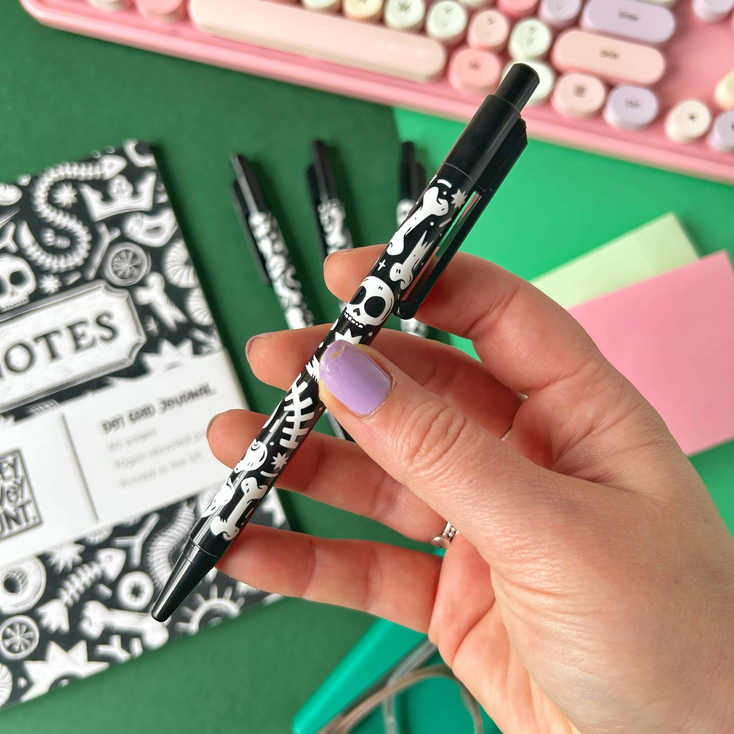 Hand holding a black and white skull and bones patterned pen with stationery items in the background.