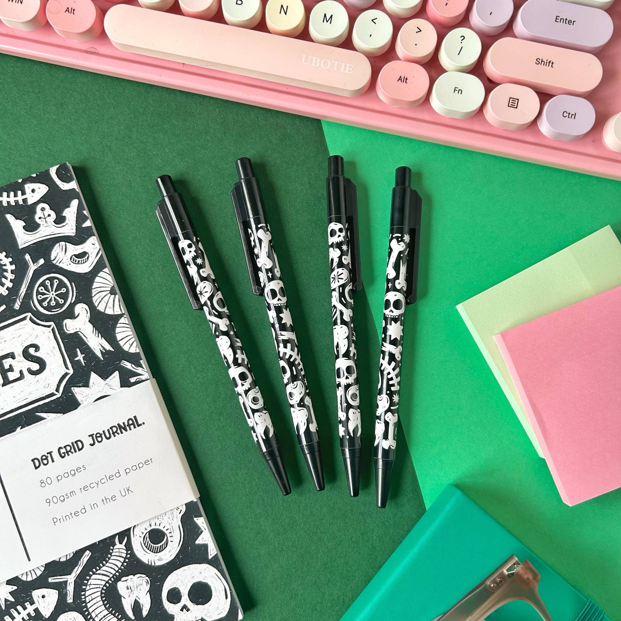 Black pens with skull design on a green surface with stationery items.