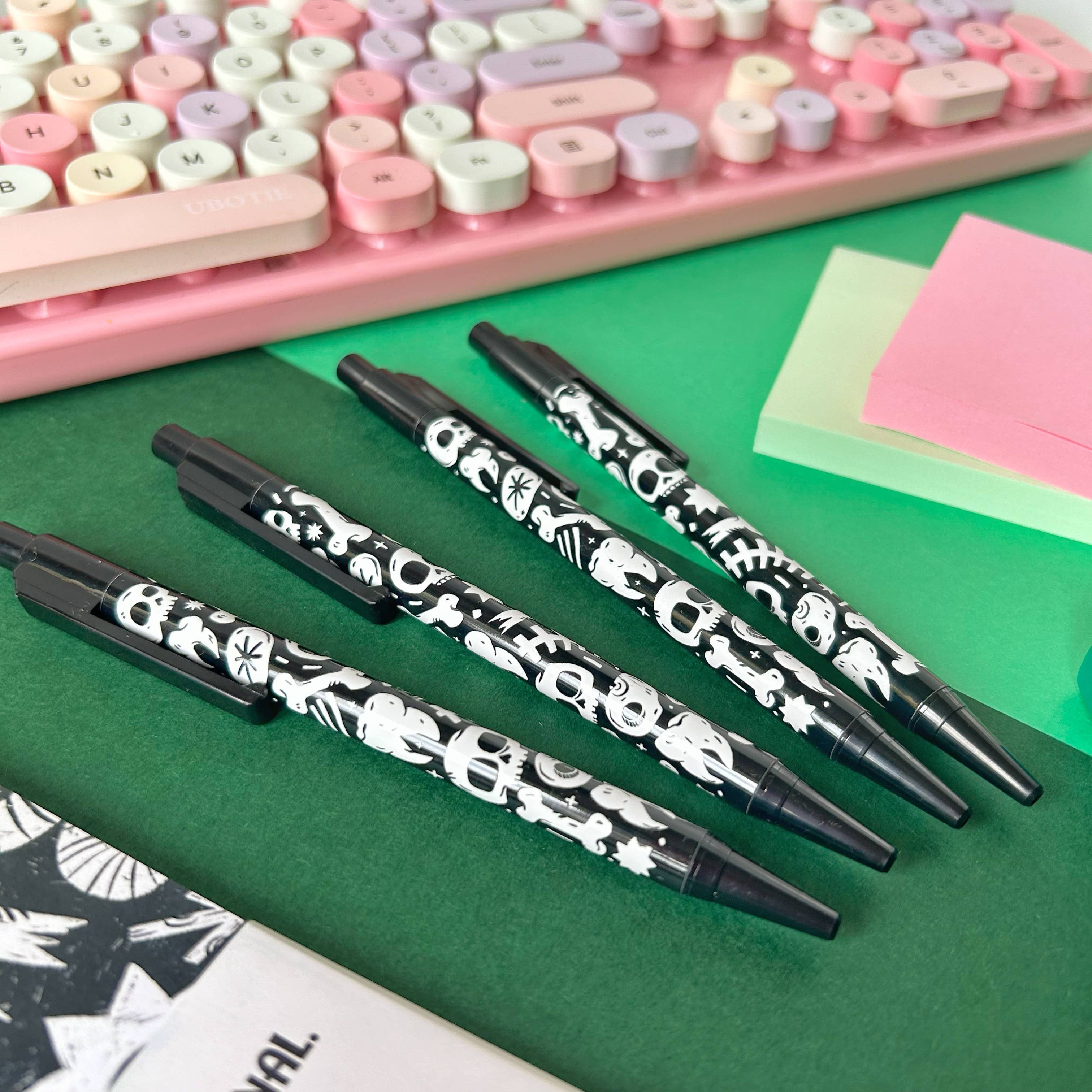 Black pens with white skull and bones patterns on a green surface with a pink keyboard in the background.