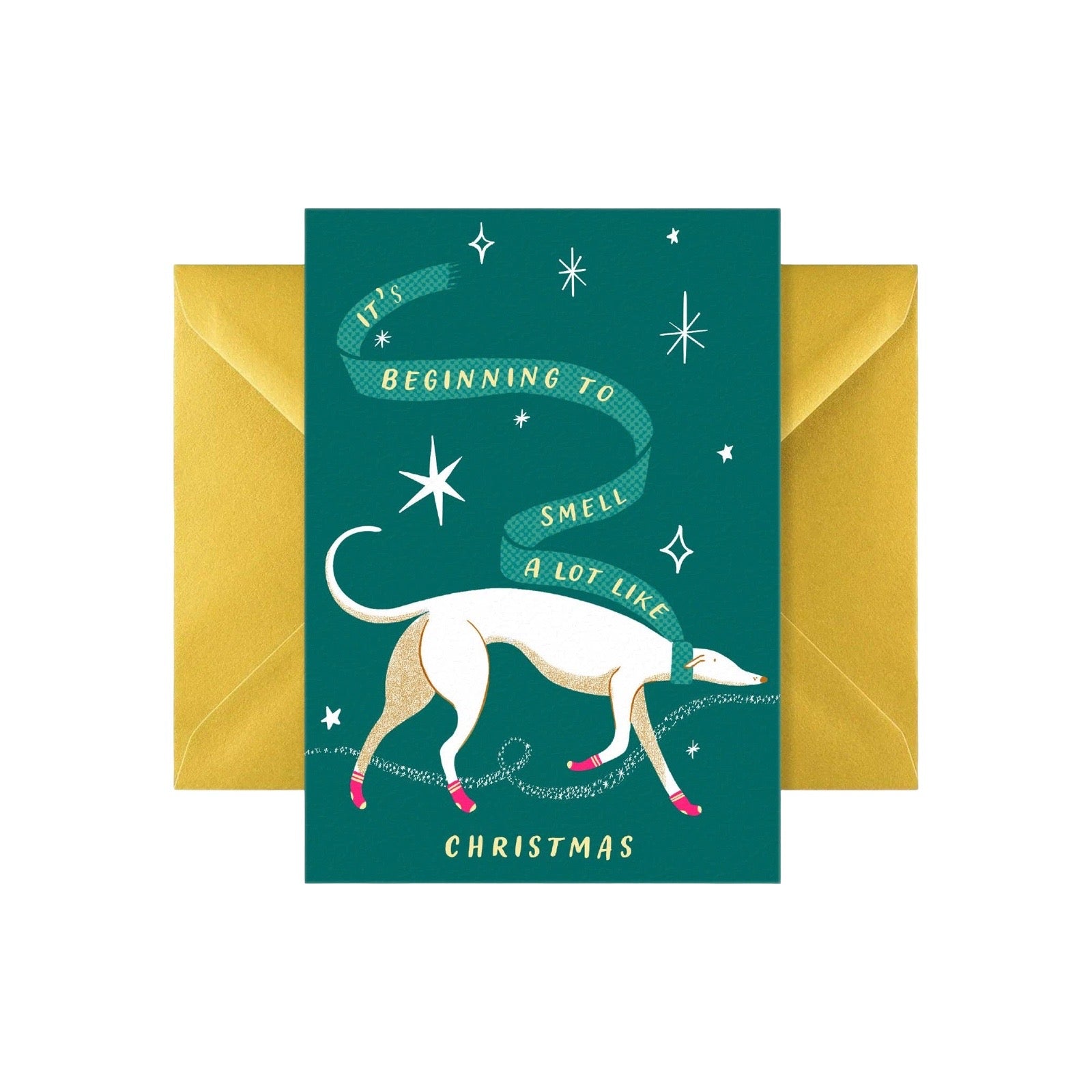 A Christmas greeting card with a dark teal background, featuring a white and beige dog with pink shoes under a green ribbon that reads 'It's Beginning to Smell a Lot Like Christmas'. The card is displayed in front of a gold envelope on a white background.
