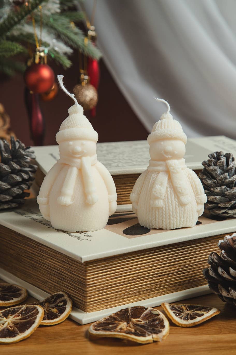 Two cream-colored snowman candles styled on top of vintage books, surrounded by pinecones and dried orange slices, with a blurred Christmas tree in the background.