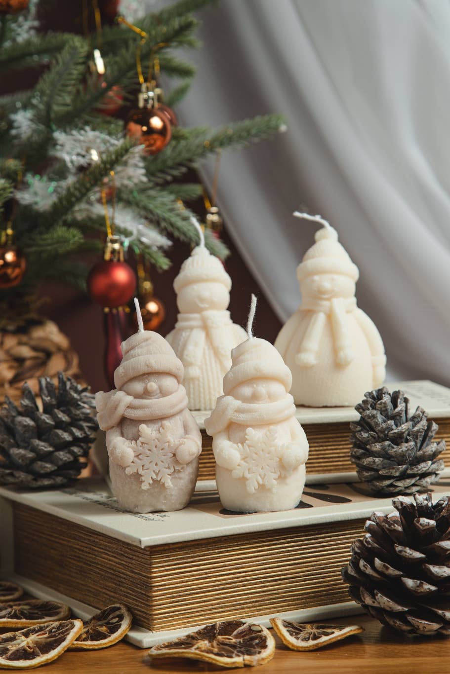Four snowman-shaped candles in cream and light beige tones, styled on a wooden surface with pinecones, dried orange slices, and a decorated Christmas tree in the background.