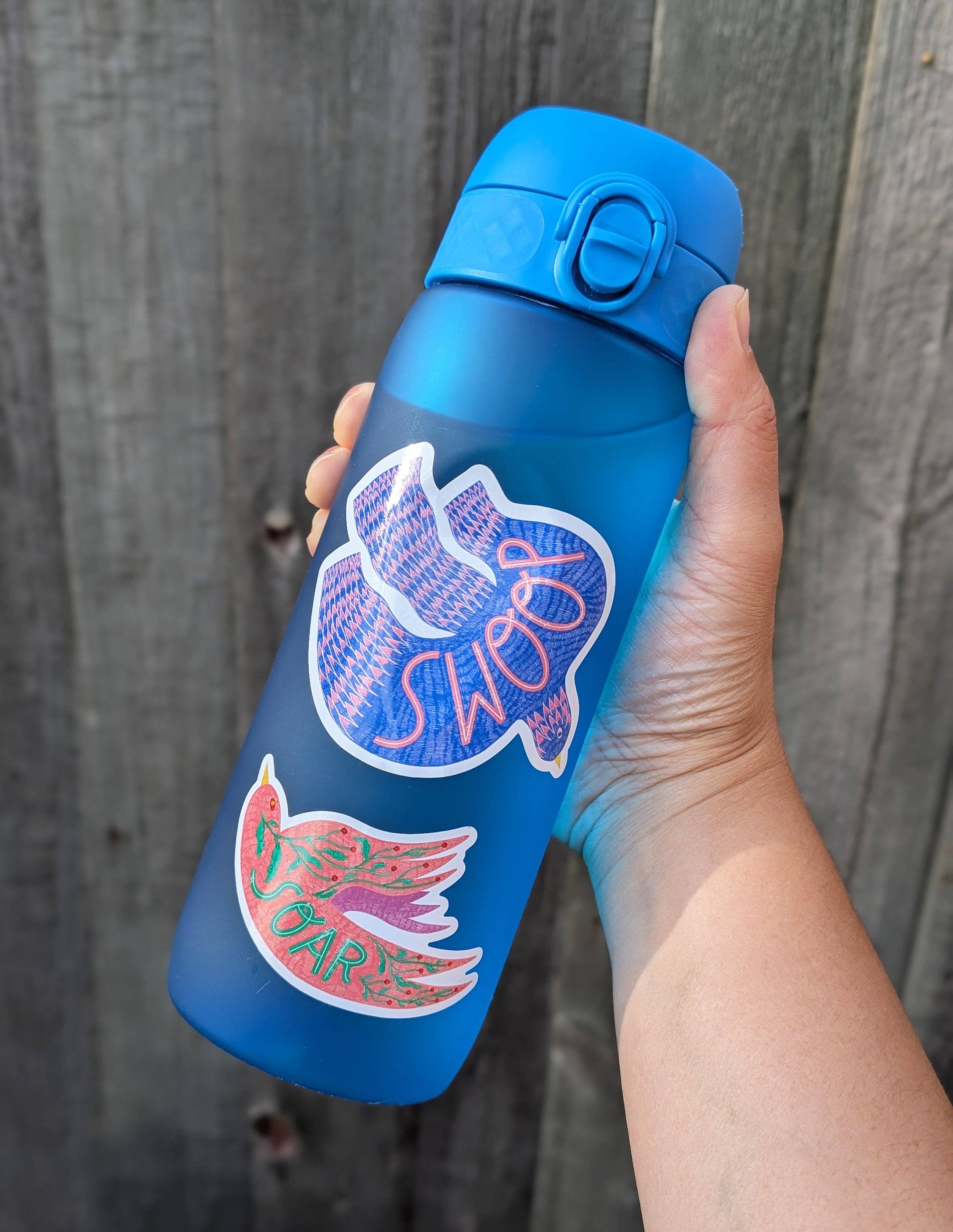 Blue water bottle with stickers held by a hand against a wooden background.