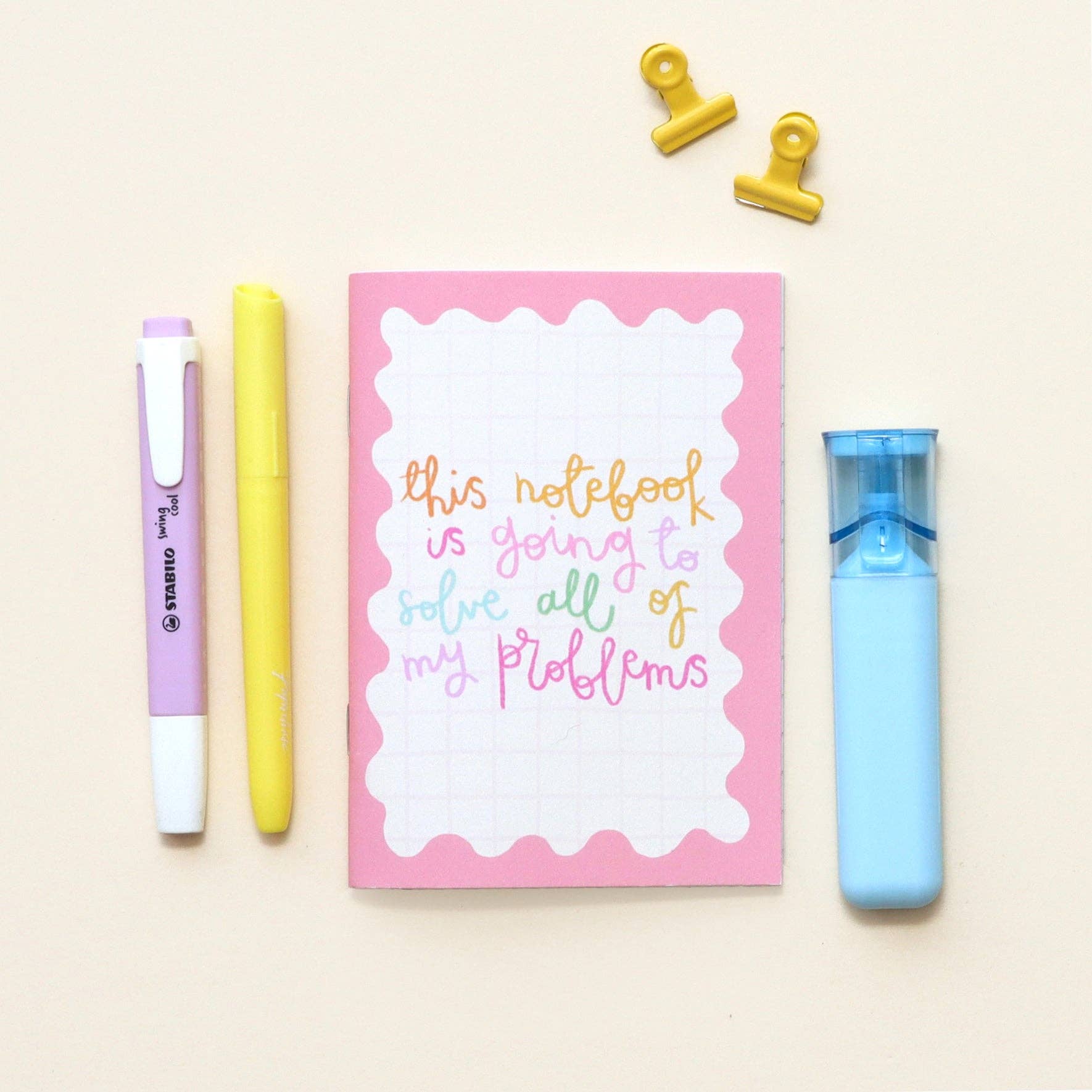Pink notebook with colourful text, three markers, and clips on a beige background.