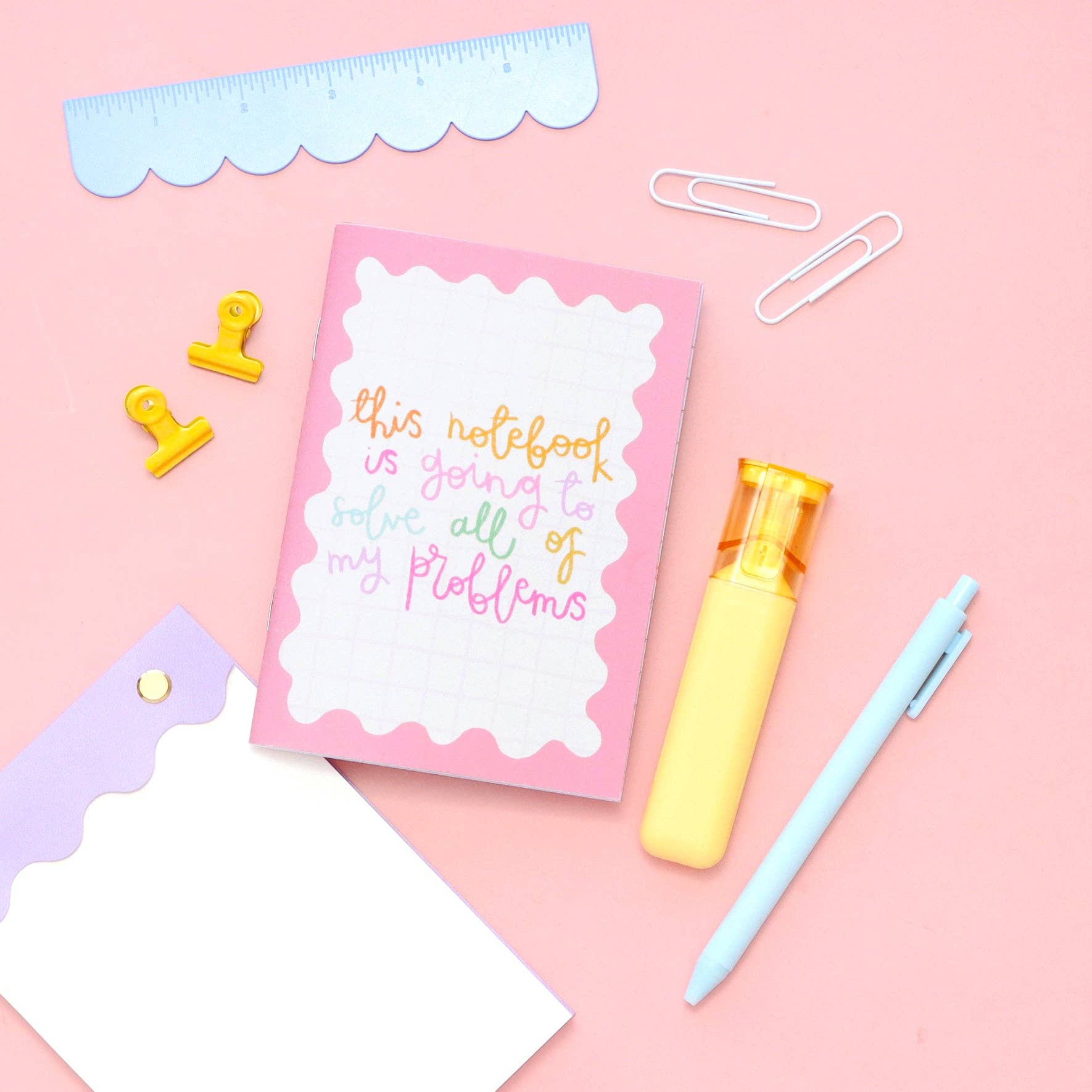 Pink notebook with text, yellow pen, blue pen, and stationery items on a pink background.