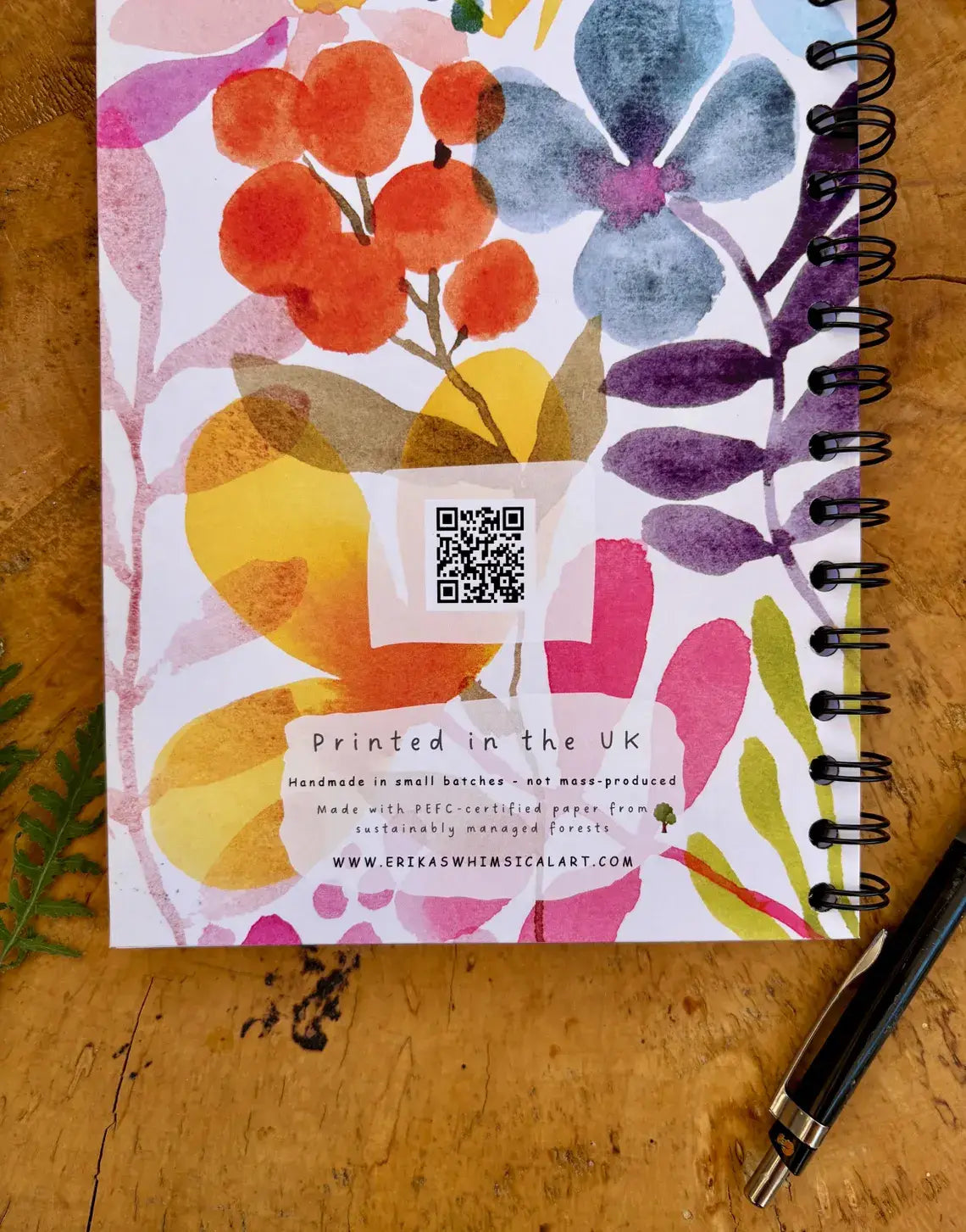 A spiral-bound notebook with a colorful watercolor floral cover lies on a wooden tabletop next to a black pen. The cover features orange berries, blue and purple flowers, and yellow leaves on a white background, with text reading 'Printed in the UK' and a QR code.