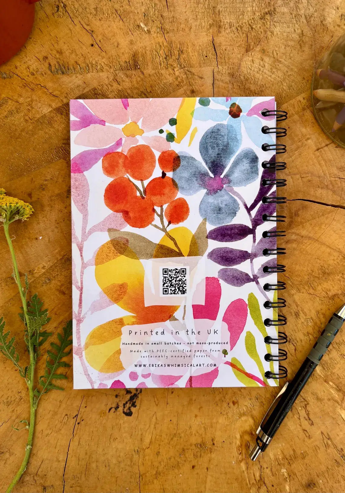A spiral-bound notebook with a colorful watercolor floral cover lies on a rustic wooden tabletop, alongside a black pen and small yellow flowers. The cover features a white base with pink, orange, blue, yellow, and purple watercolor flowers and leaves, plus a QR code and printed text near the bottom.
