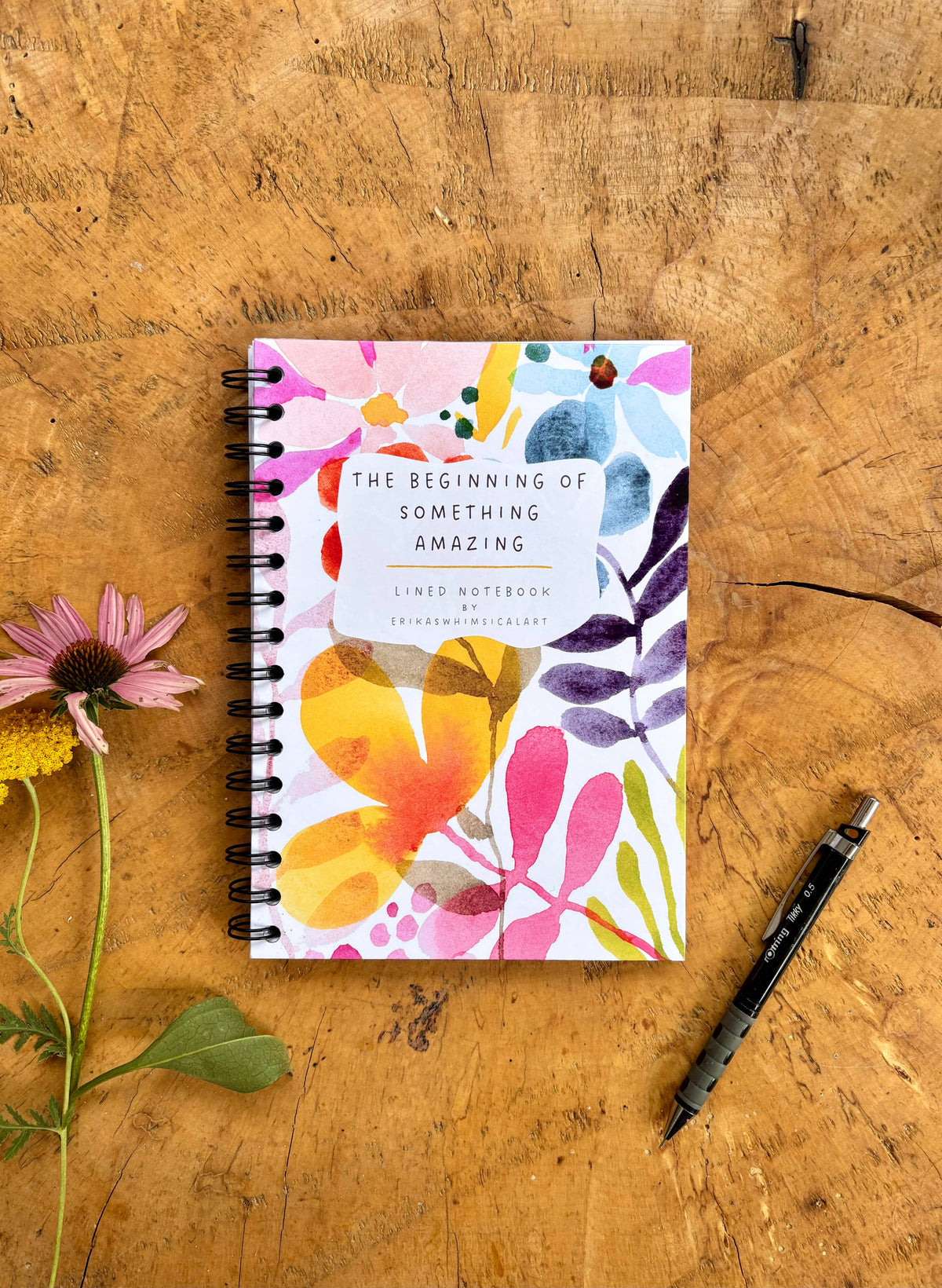 A spiral-bound notebook with a colorful watercolor floral cover lies on a rustic wooden surface, styled with a pink flower on the left and a black pen on the right. The notebook cover reads, 'THE BEGINNING OF SOMETHING AMAZING'.