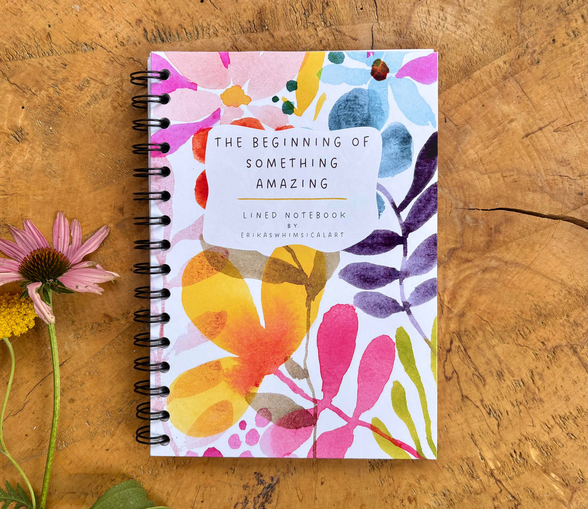 A spiral-bound lined notebook with a colorful floral cover sits on a rustic wooden surface next to pink and yellow flowers. The cover features the text 'THE BEGINNING OF SOMETHING AMAZING'.