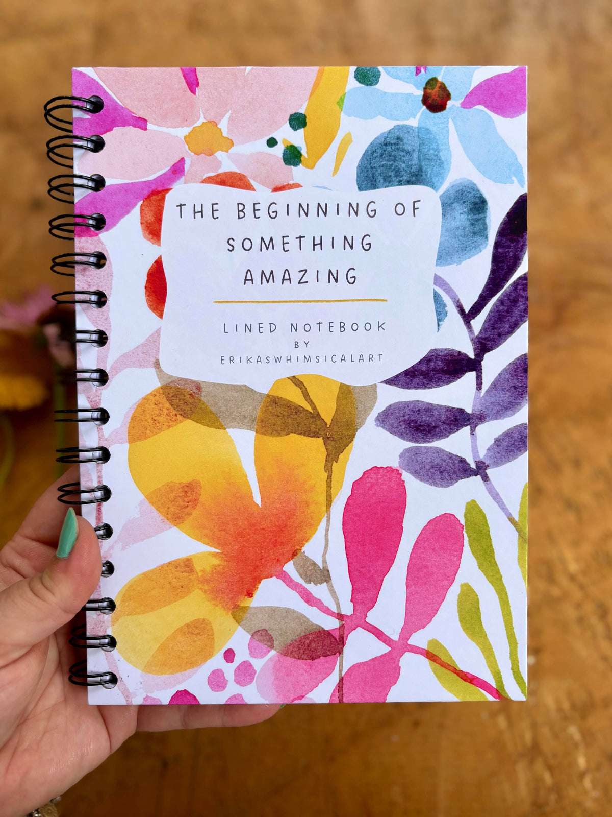 A hand holds a spiral-bound lined notebook with a colorful watercolor floral cover. The cover features the text 'THE BEGINNING OF SOMETHING AMAZING' and 'ERIKASWHIMSICALART'.