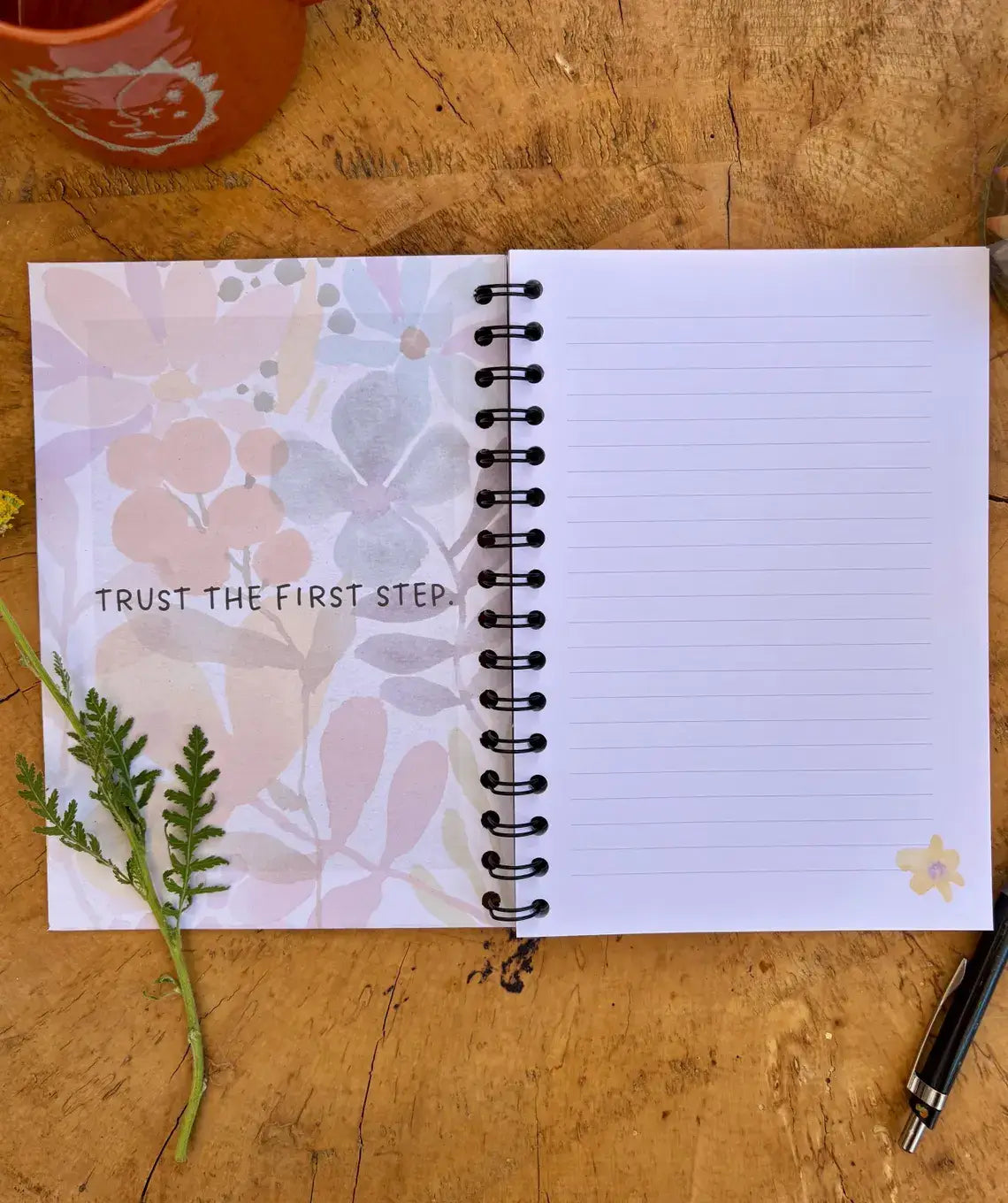 An open spiral-bound lined notebook lies on a rustic wooden surface. The left page has a soft pastel floral pattern with the words 'TRUST THE FIRST STEP.
