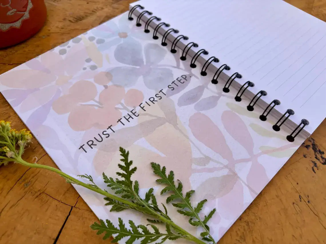 A spiral-bound notebook with a white cover featuring a soft pastel floral pattern and the printed phrase 'TRUST THE FIRST STEP,' photographed on a wooden surface with a green stem placed across the lower part of the page.