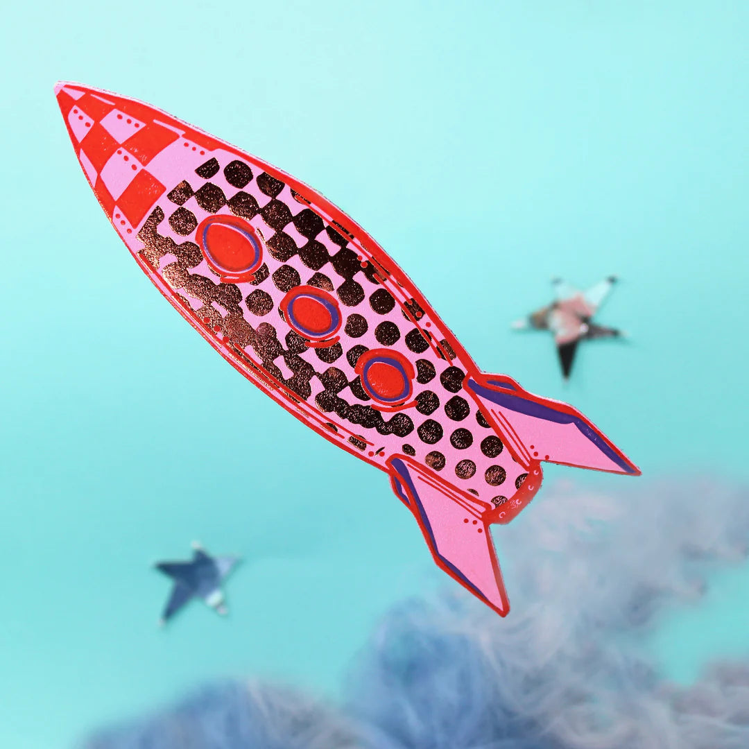 Pink rocket bookmark with black and red details against a blue sky.