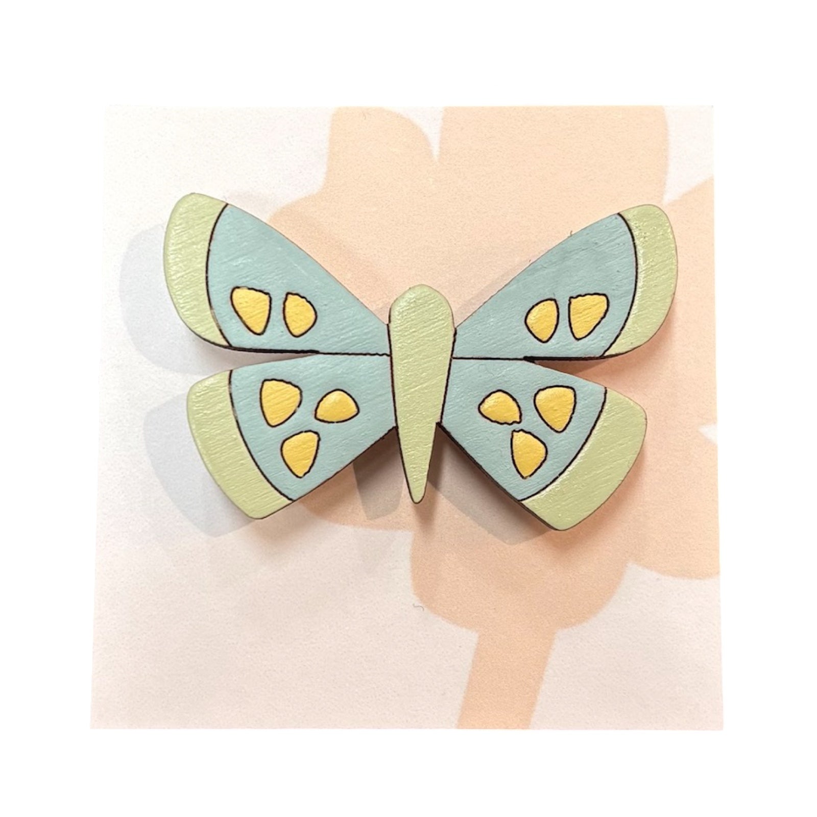 A speckled butterfly wooden brooch, hand-painted with colourful wings in pale blue and pale green with yellow spots on a white backing card.