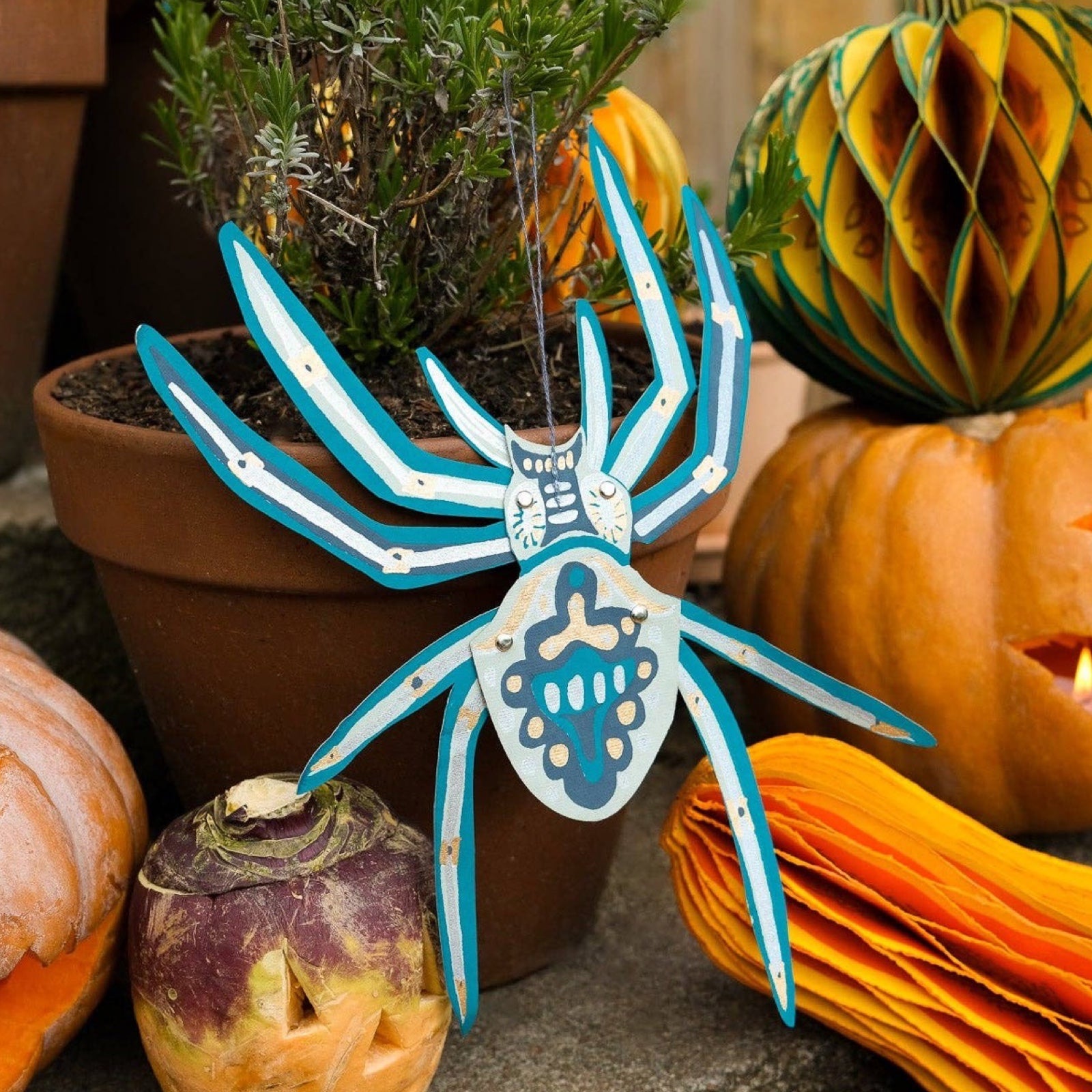 A paper hanging spider decoration hanging from a plant next to a pumpkin.