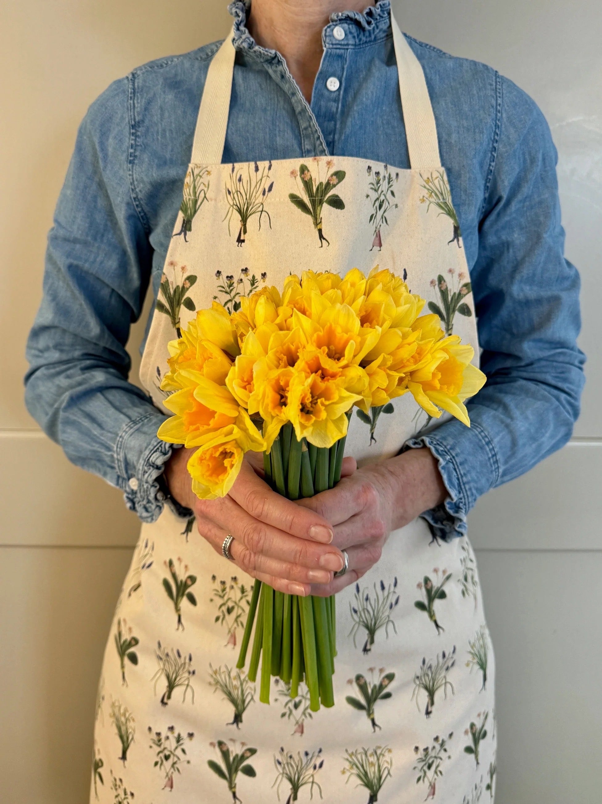 Person wearing a floral apron holding a bouquet of yellow flowers against a neutral background.