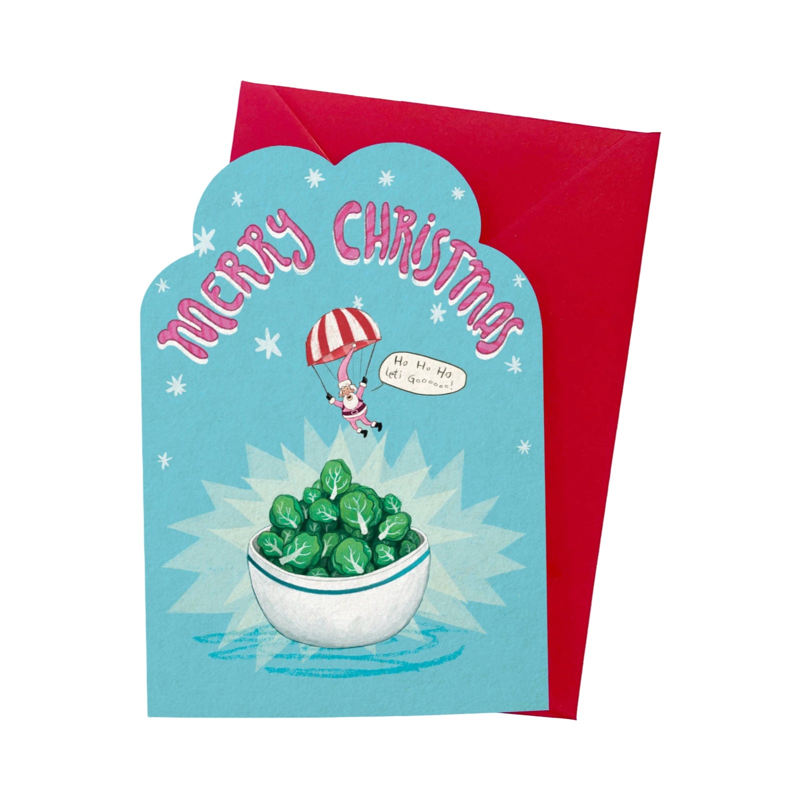 A Christmas greeting card with a scalloped light blue front featuring the text 'Merry Christmas' in pink, an illustration of a Santa Claus figure under a red-and-white striped parachute above a bowl of green sprouts, with a red envelope behind it on a white background.