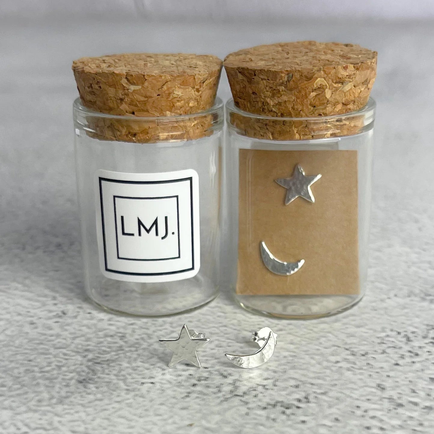 Two glass jars with cork lids, one labeled 'LMJ.' and the other with star and moon earings on brown card, on a textured surface. A star and moon earring are sat in front of the jars.