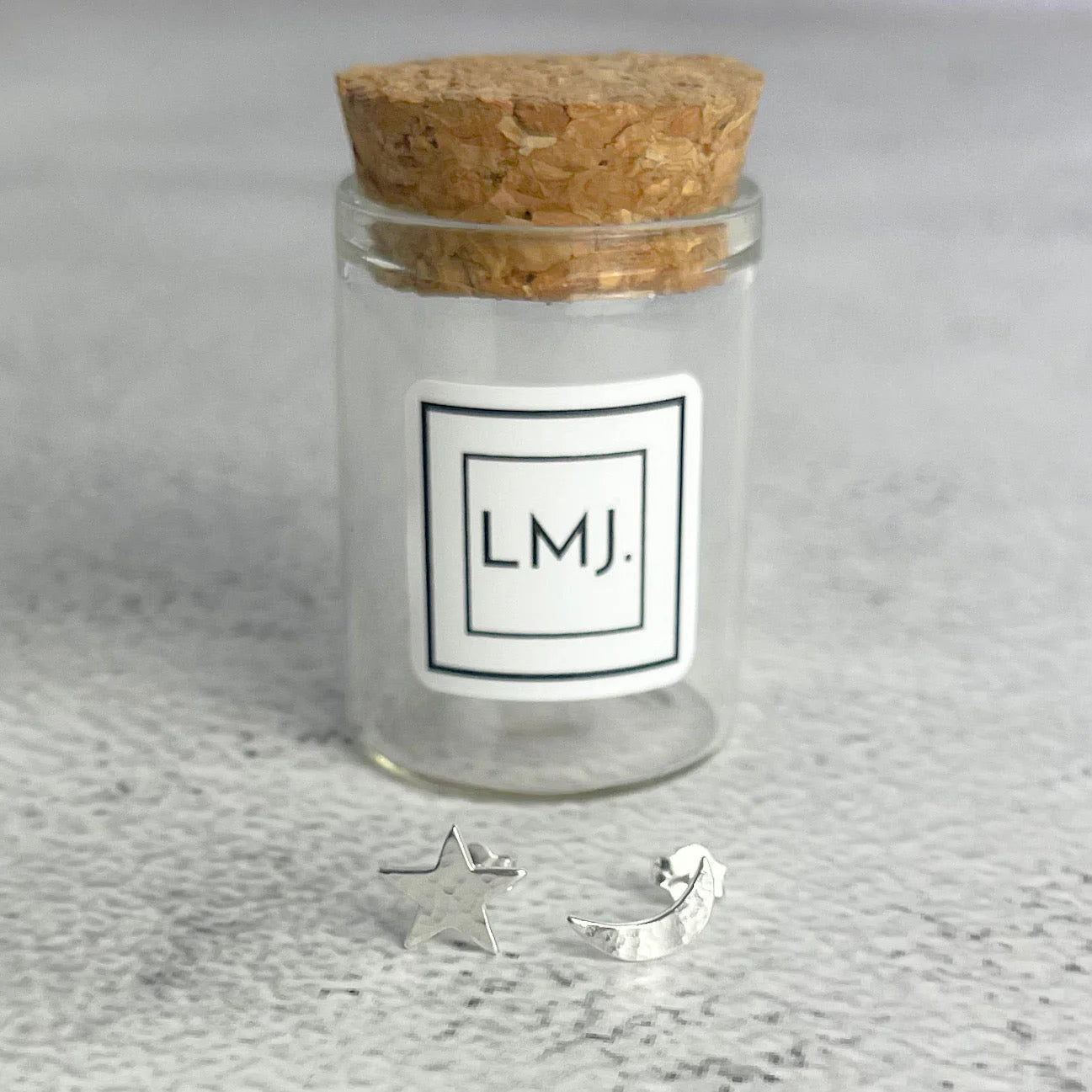 Silver star and moon earrings in front of a glass jar with cork lid labeled 'LMJ'.