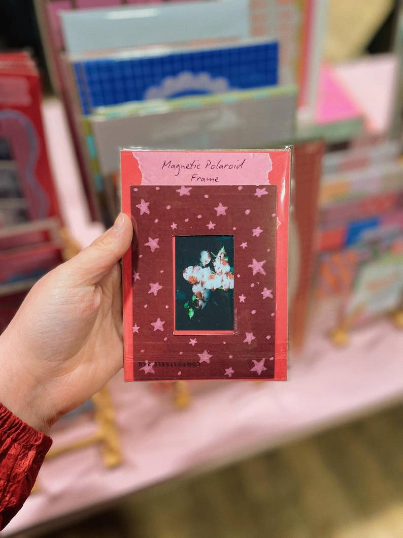A hand holding a packaged magnetic polaroid frame with a dark red star-patterned border and a central black window showing a flower photo.