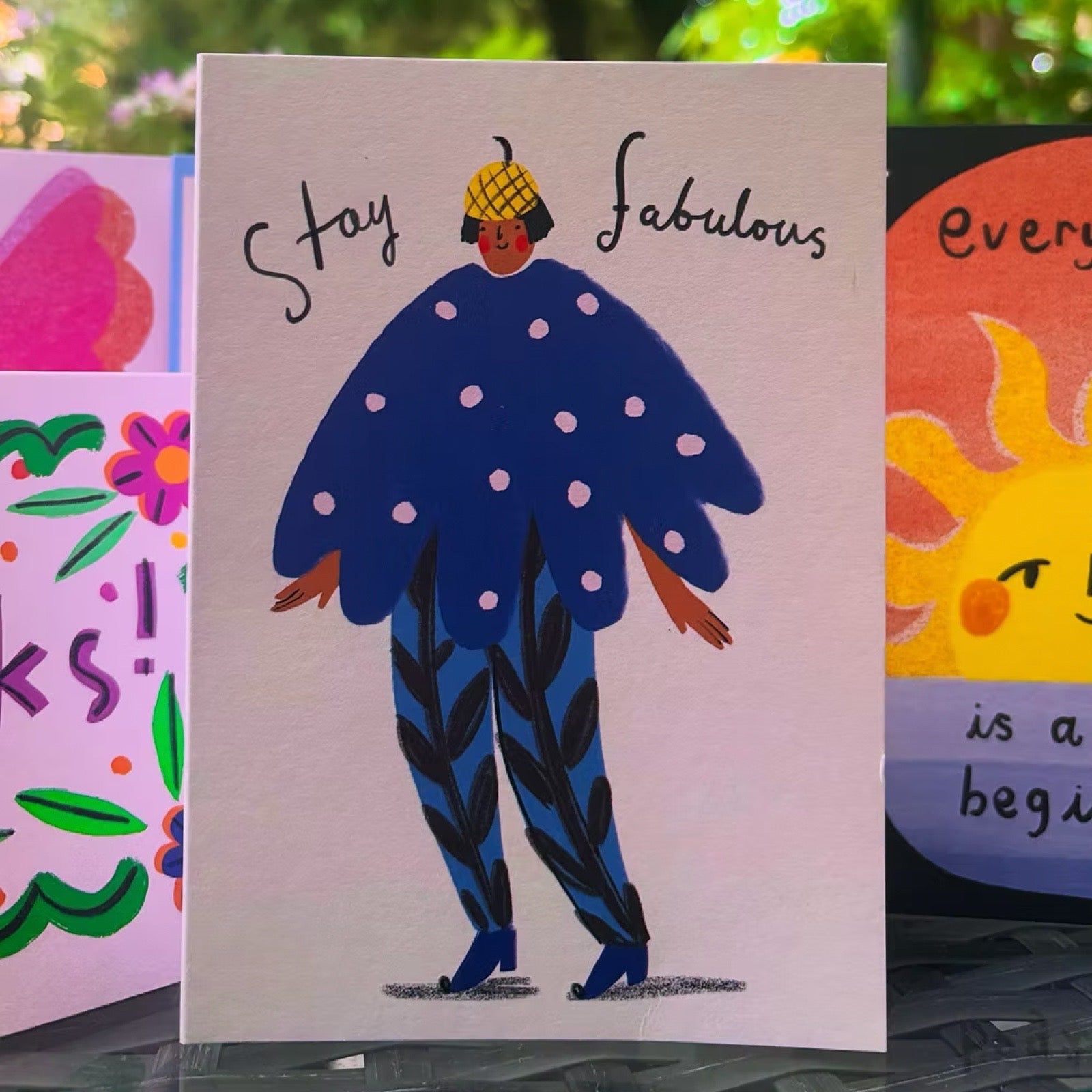 A square greeting card featuring a whimsical illustration of a person with a yellow checkered hat, a blue polka-dot cape, and patterned blue pants, with the words 'Stay fabulous' written above. The card is displayed on a glass surface with other colourful cards partially visible in the background.