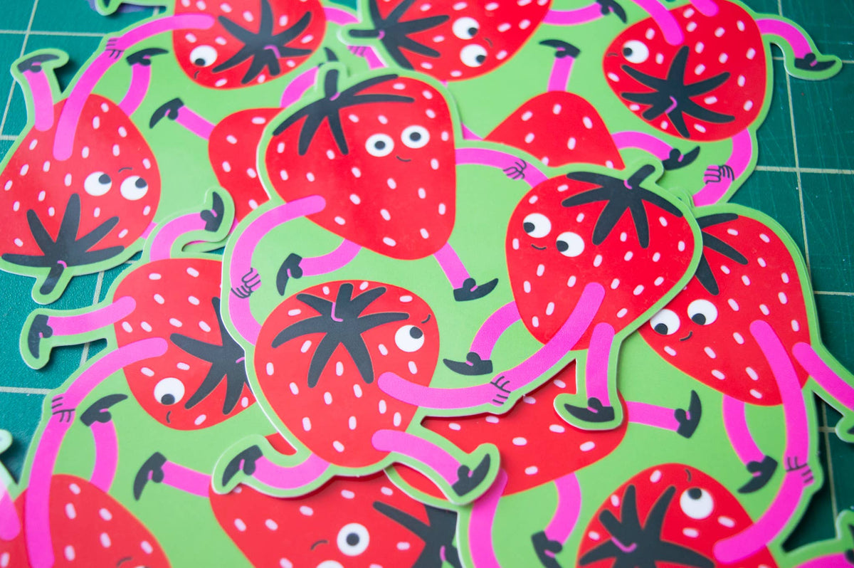 A pile of die-cut strawberry stickers with cartoon eyes, arms, and legs, displayed on a green cutting mat.
