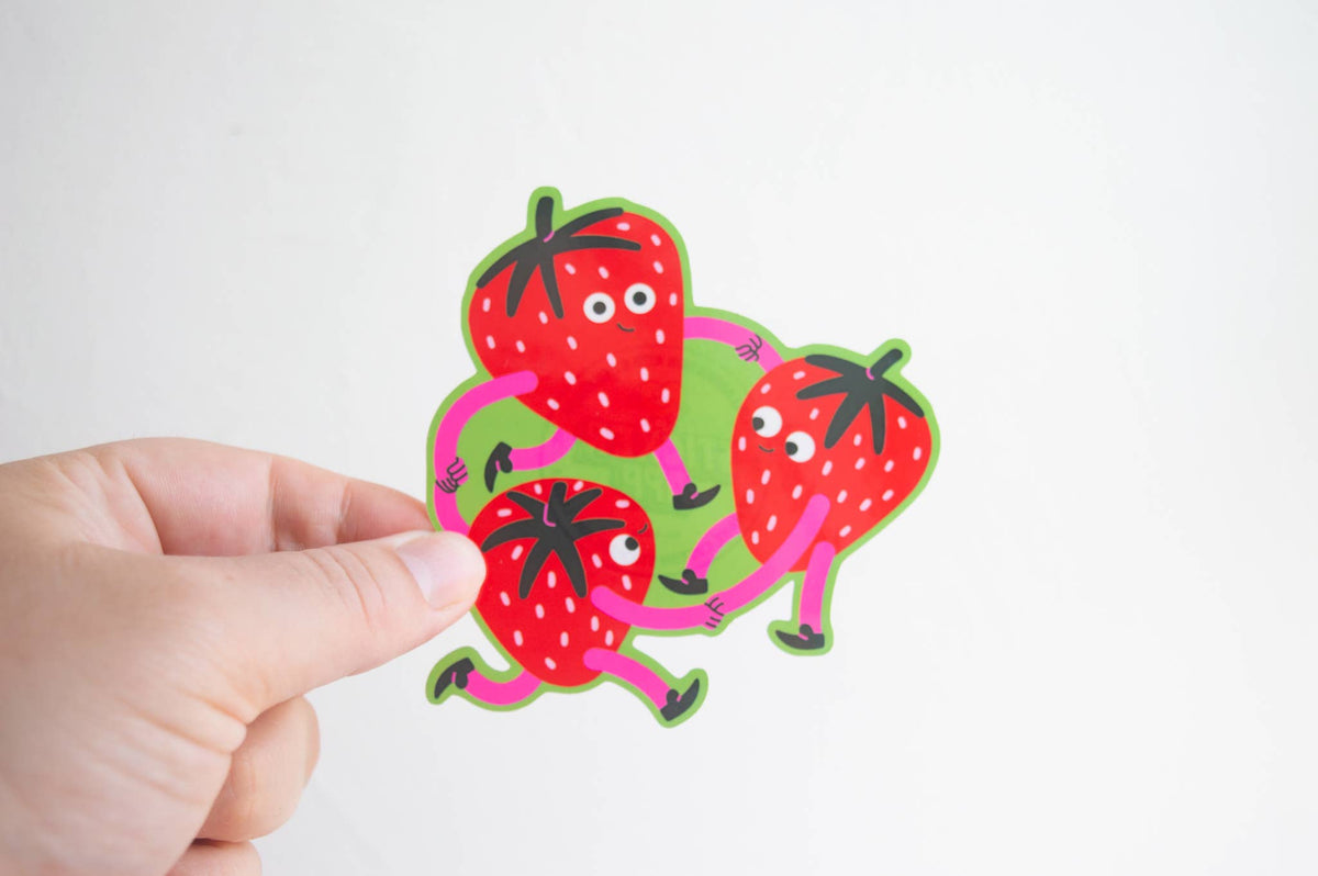 A hand holding a die-cut sticker featuring three cartoon strawberries with pink limbs and white eyes, set against a plain white background.