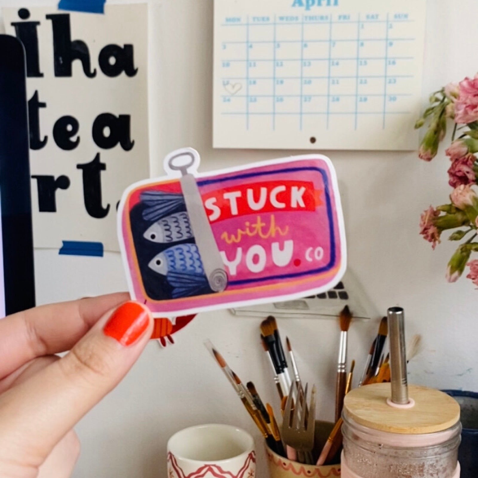Hand holding a decorative sticker with text and fish design in an indoor setting.