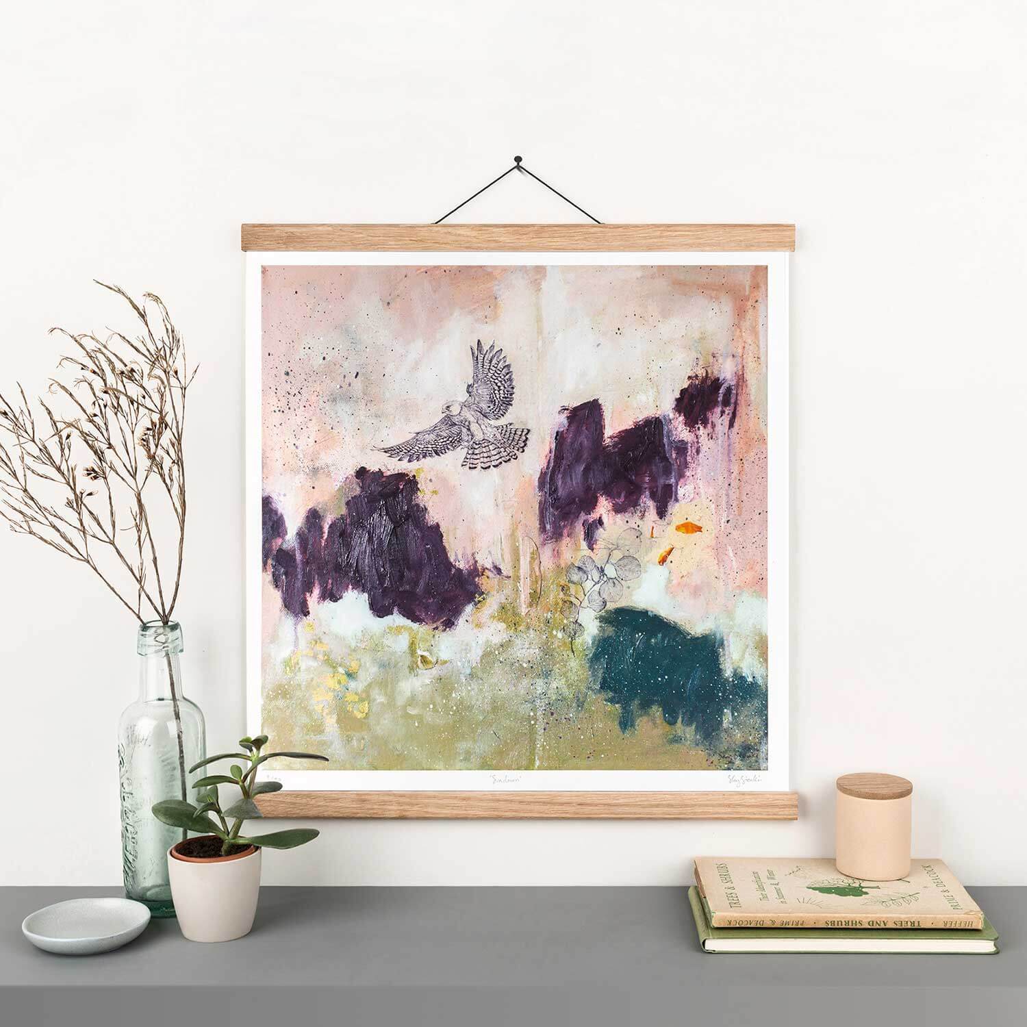 A framed bird print featuring an abstract background with purple, pink, and green splashes of color, displayed on a white wall above a plant pot.