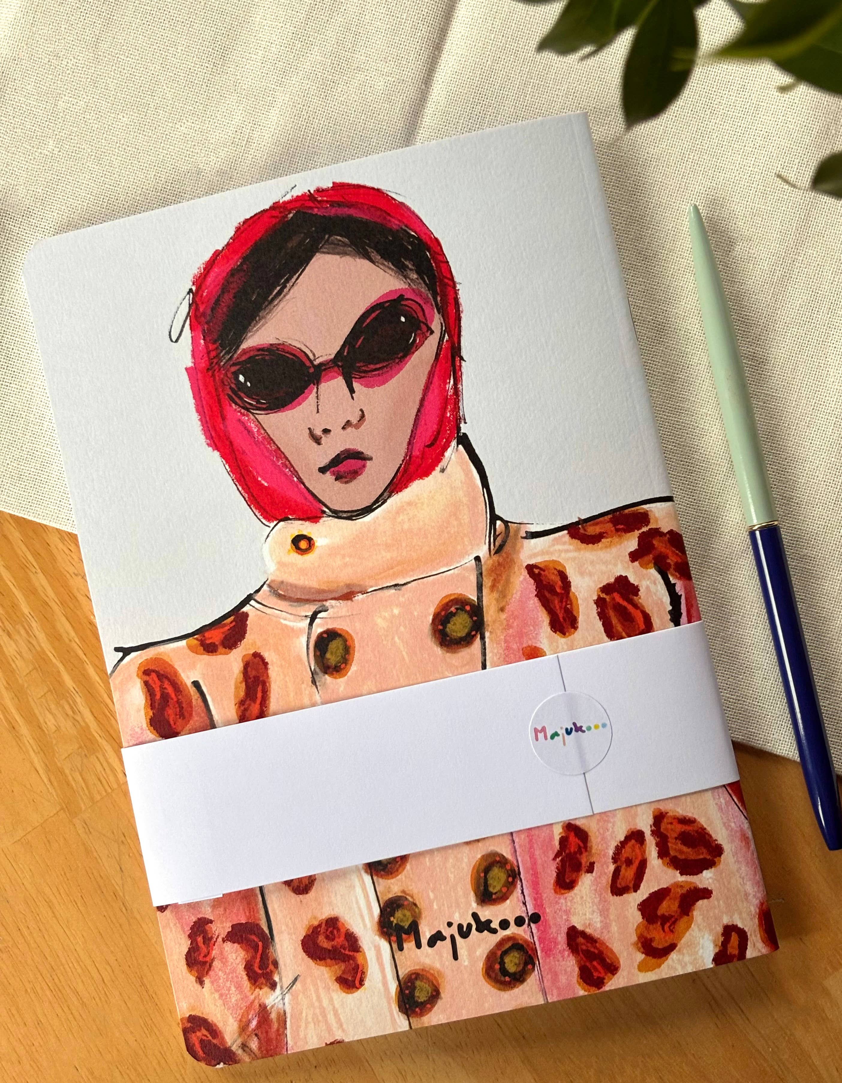 Notebook with a colorful illustration of a person wearing a red headscarf and sunglasses, placed on a wooden surface.