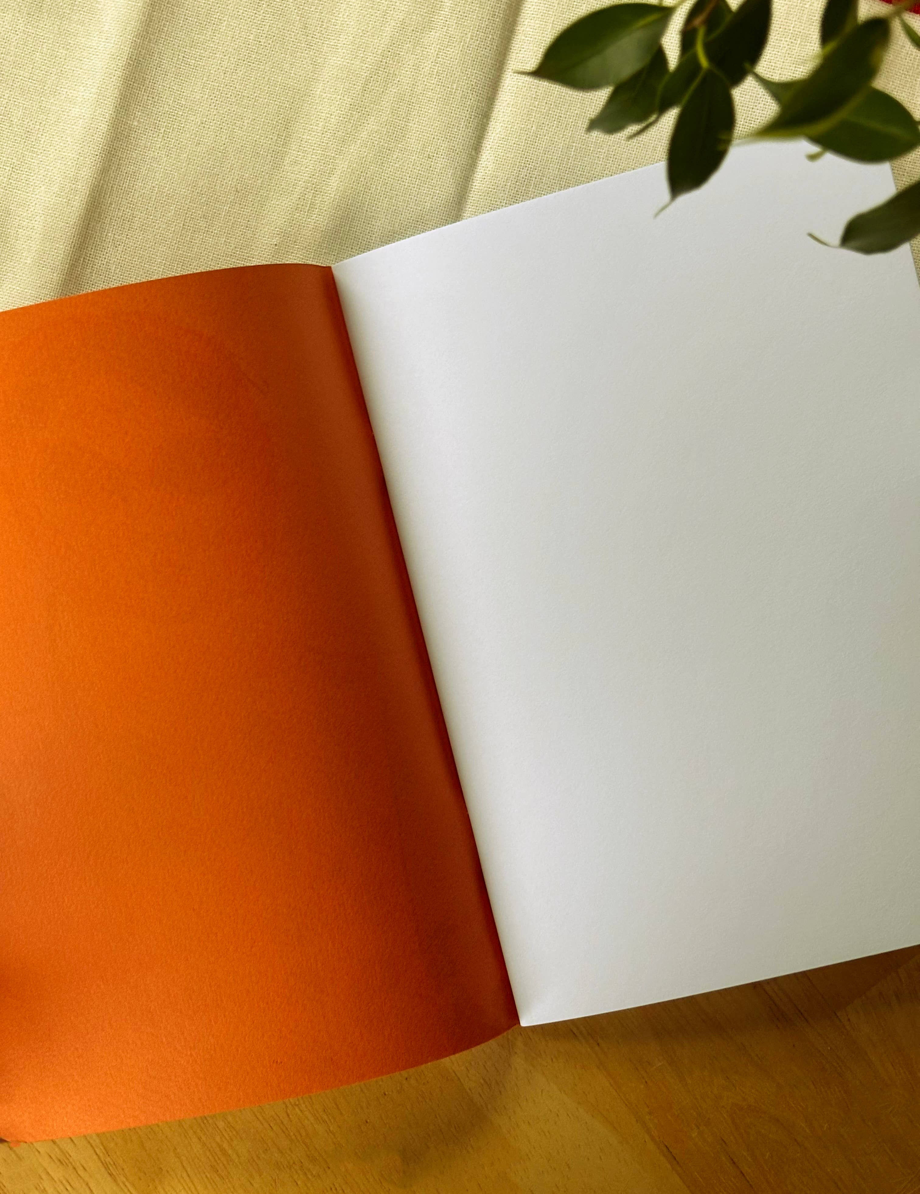 An open notebook with an orange side and a white page, on a wooden surface with green leaves in the background.