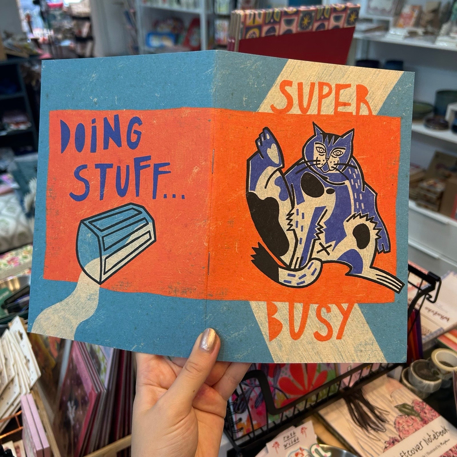 A hand holds a notebook featuring a colourful illustration of a busy cat and the text 'DOING STUFF... SUPER BUSY'.