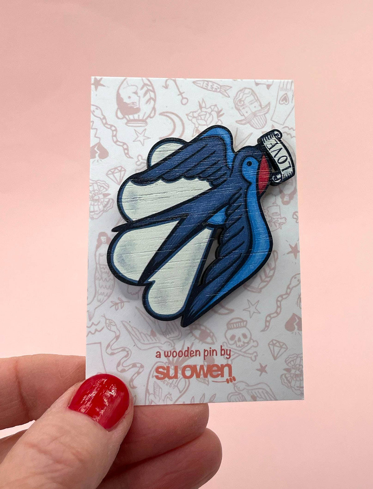 Blue bird-shaped wooden brooch on a card held by a hand with red nail polish against a pink background.