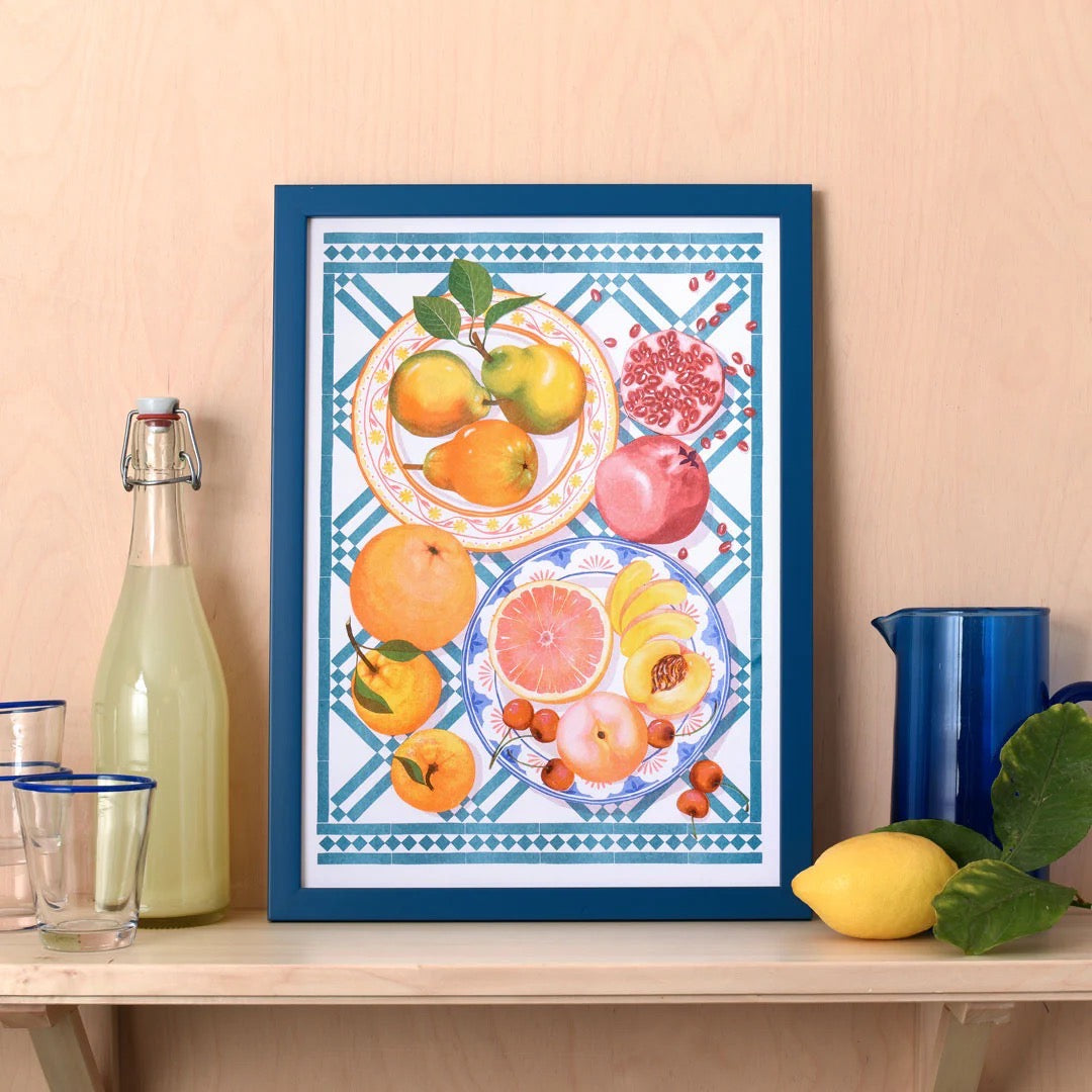 Framed artwork with fruit design on a table against a beige wall, surrounded by lemonade and lemons.