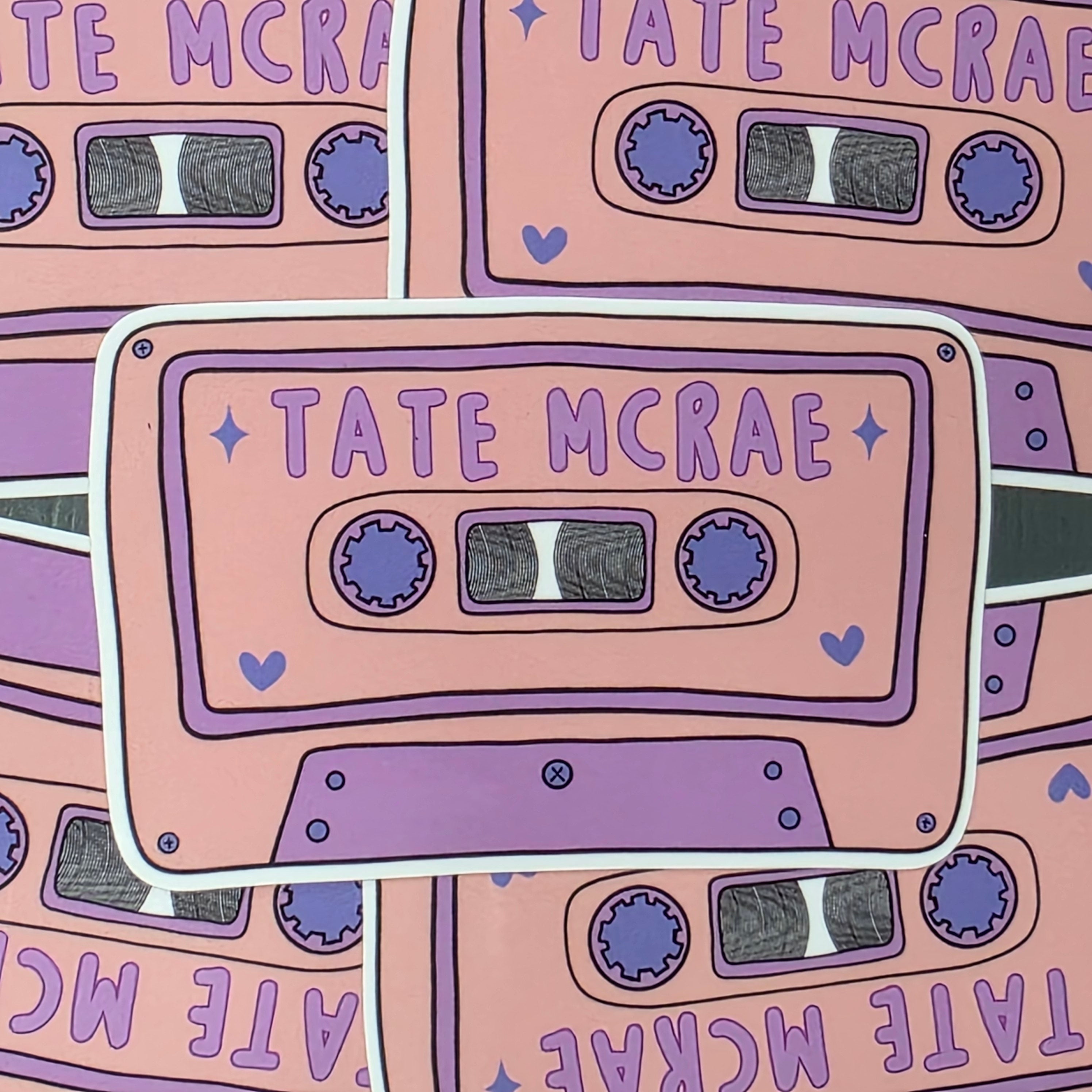 A rectangular-shaped sticker featuring an illustration of a retro cassette tape in pink and lilac with the title 'Tate McRae', decorated with purple love hearts.