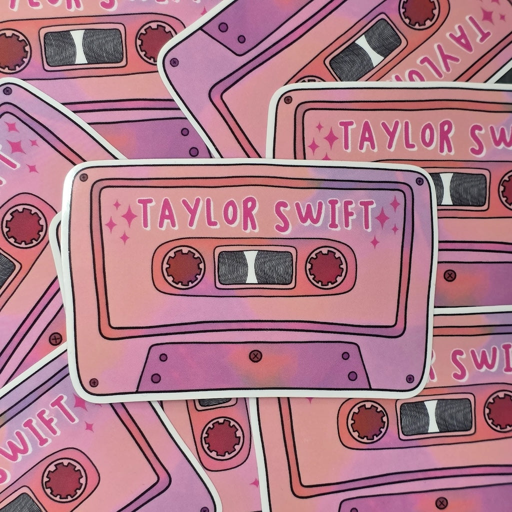 A rectangular-shaped sticker featuring an illustration of a retro cassette tape in dark and light pink with the title 'Taylor Swift'.