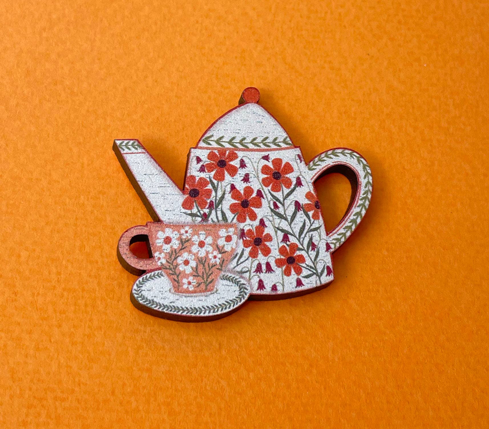A decorative wooden tea set brooch featuring a white teapot with red floral patterns, a matching teacup and saucer, and a small teapot, displayed on a textured orange background.