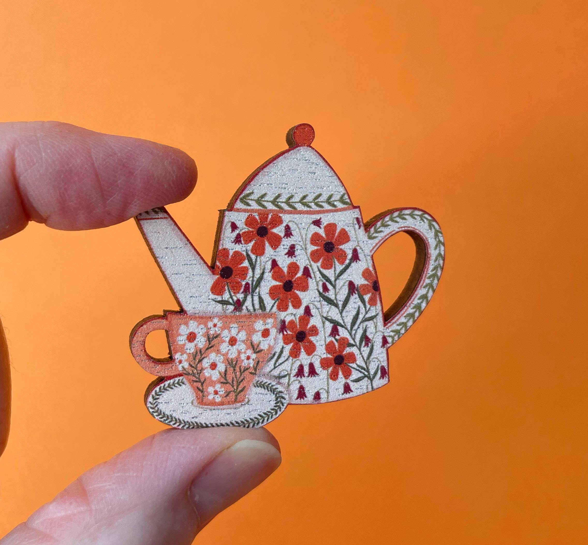 A hand holding a decorative wooden tea set brooch featuring a teapot and teacup with red flowers and green leaves, set against a solid orange background.